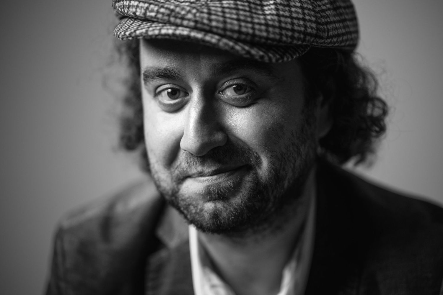 Black and white portrait of a smiling man with curly hair wearing a flat cap and blazer.