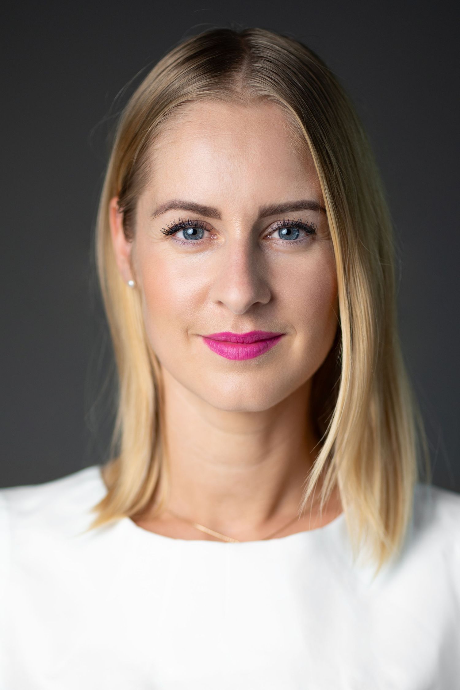 Professional headshot of a blonde woman with blue eyes and pink lipstick wearing a white top against a gray background.