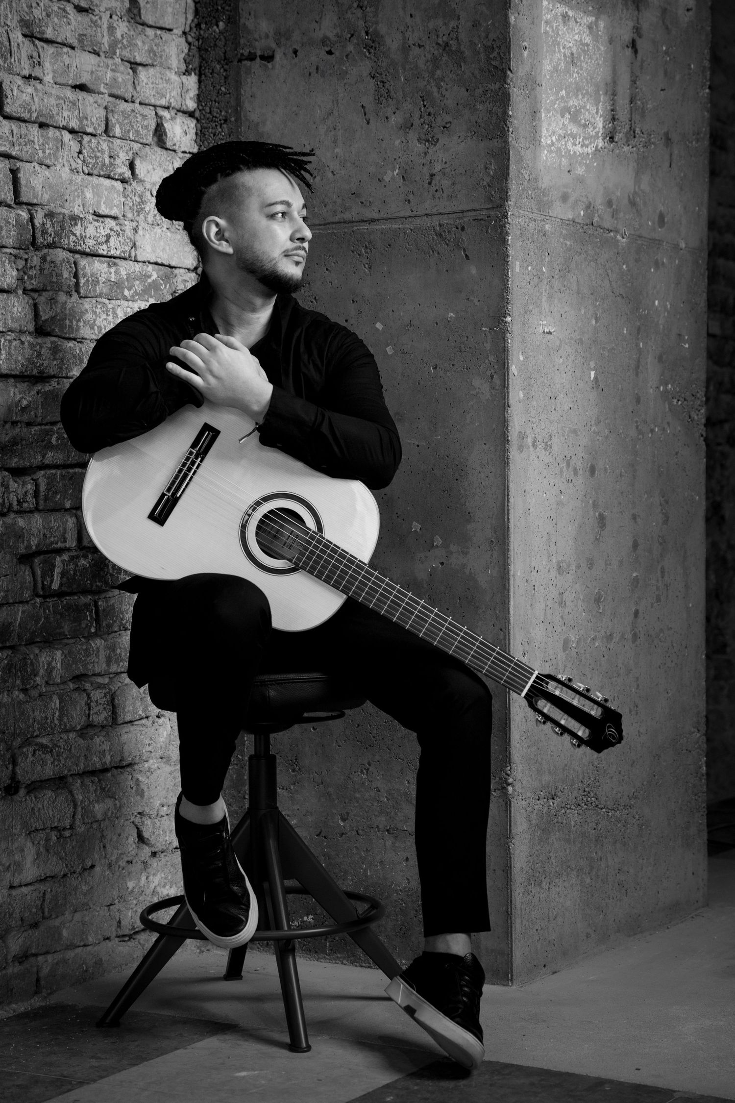 Black and white photo of a young man in a hat sitting on a stool holding an acoustic guitar against a brick wall.
