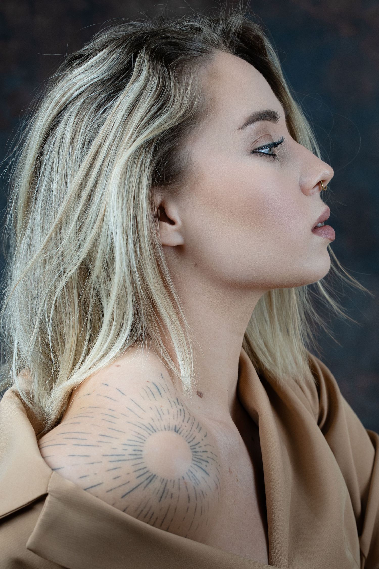 Blonde woman in profile view wearing a tan coat, showcasing a sun tattoo on her shoulder.