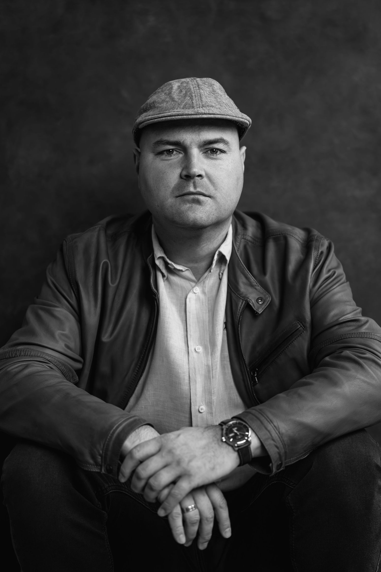 Black and white portrait of a man in a flat cap and leather jacket, sitting with hands clasped, wearing a watch.