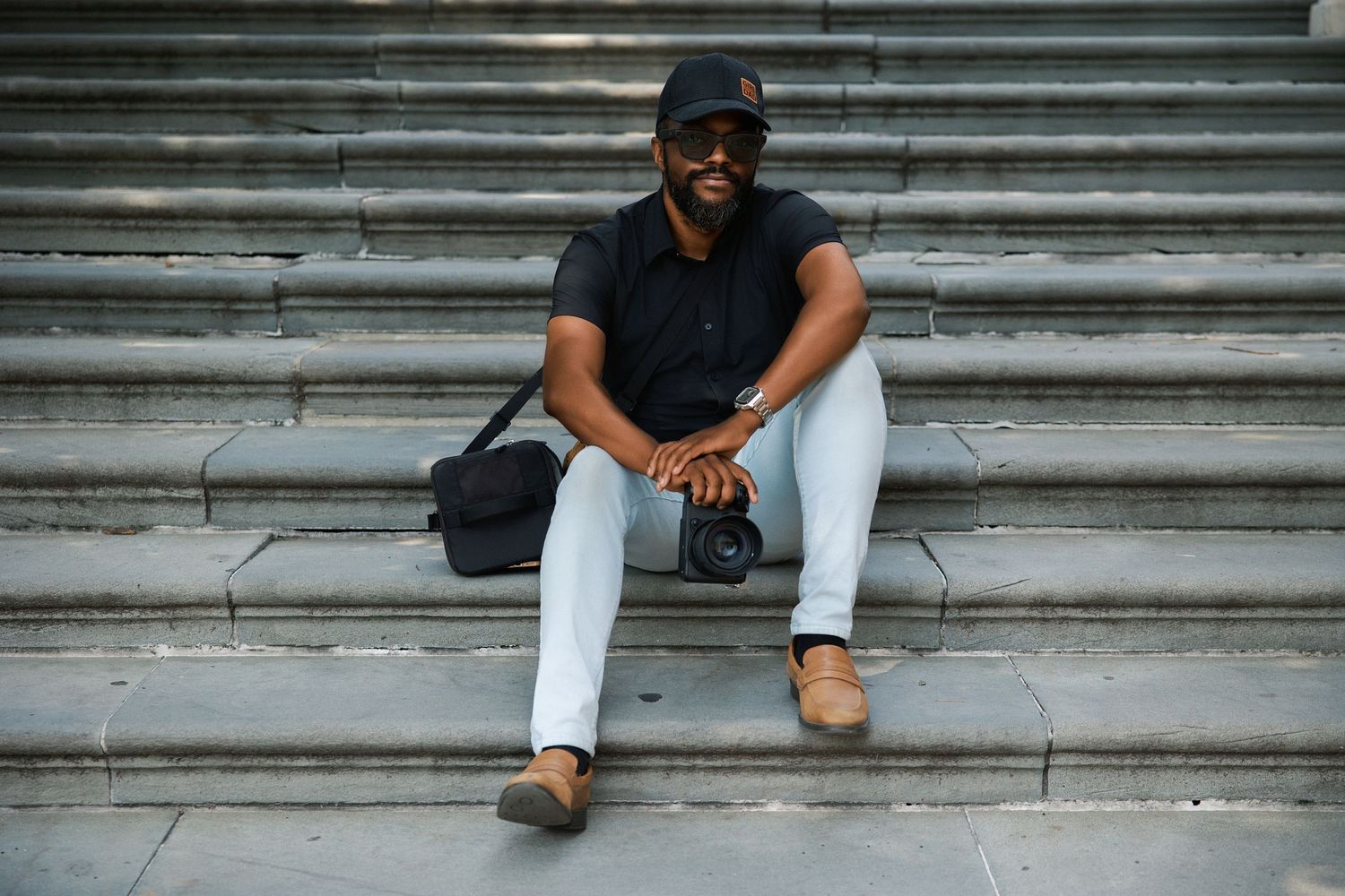A smiling Black man in a black shirt and white pants sits on stone steps holding a camera with a bag beside him.