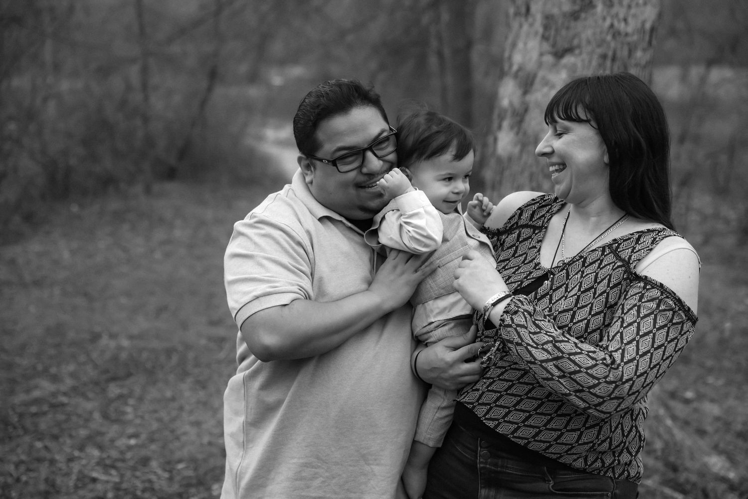 A heartwarming black and white family portrait showing parents joyfully embracing their baby in an outdoor setting.  L Visions Photography.