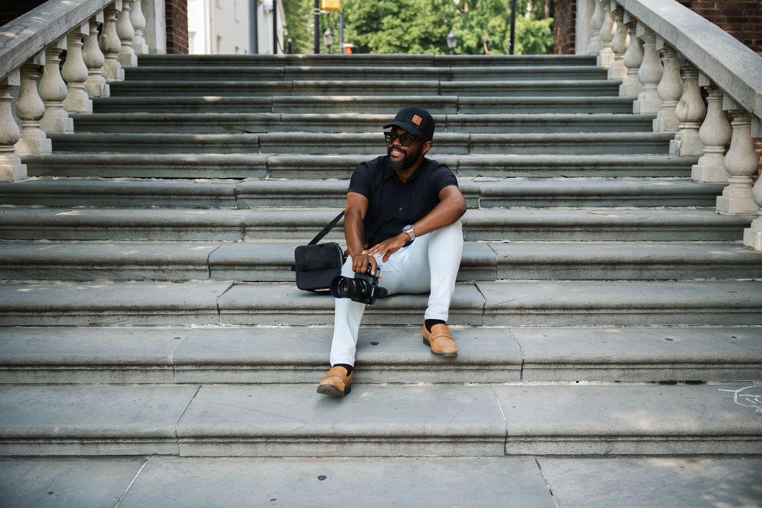 Man in black outfit and white pants sitting on stone steps with a camera bag, urban architecture background.