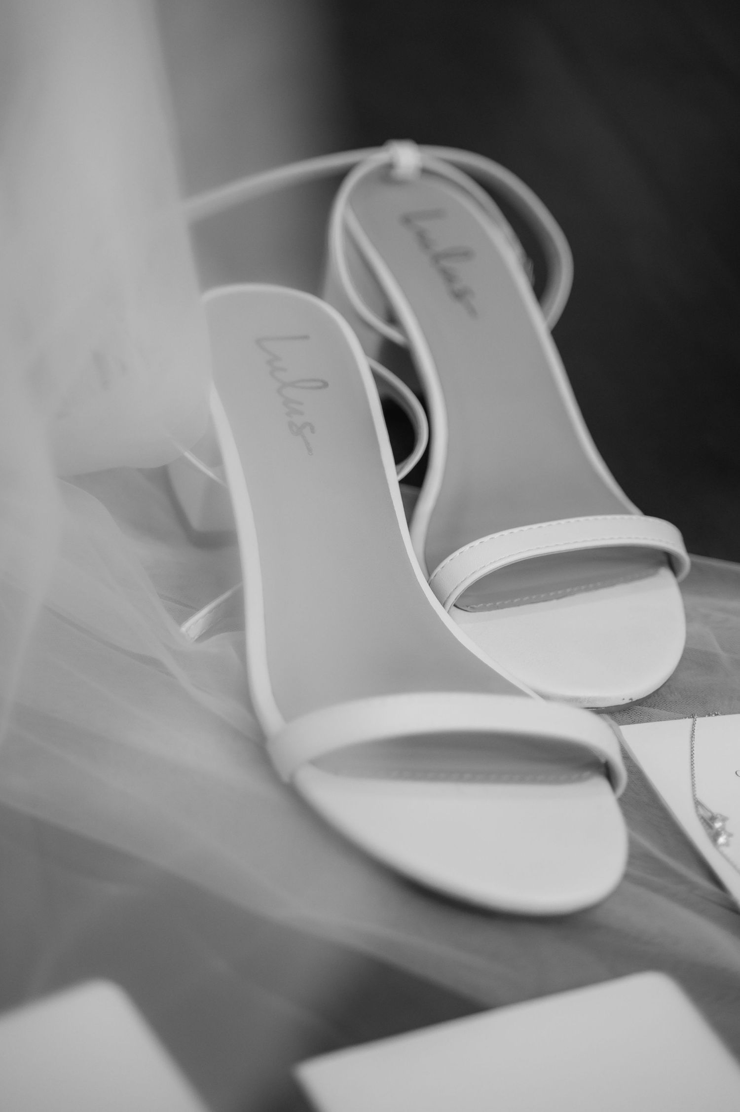 Elegant white bridal sandals displayed on white fabric in black and white photography.