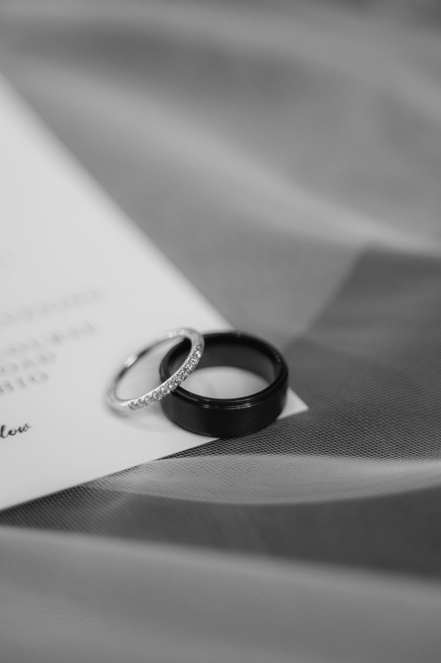 Two wedding rings, one black and one diamond, resting on paper in monochrome.