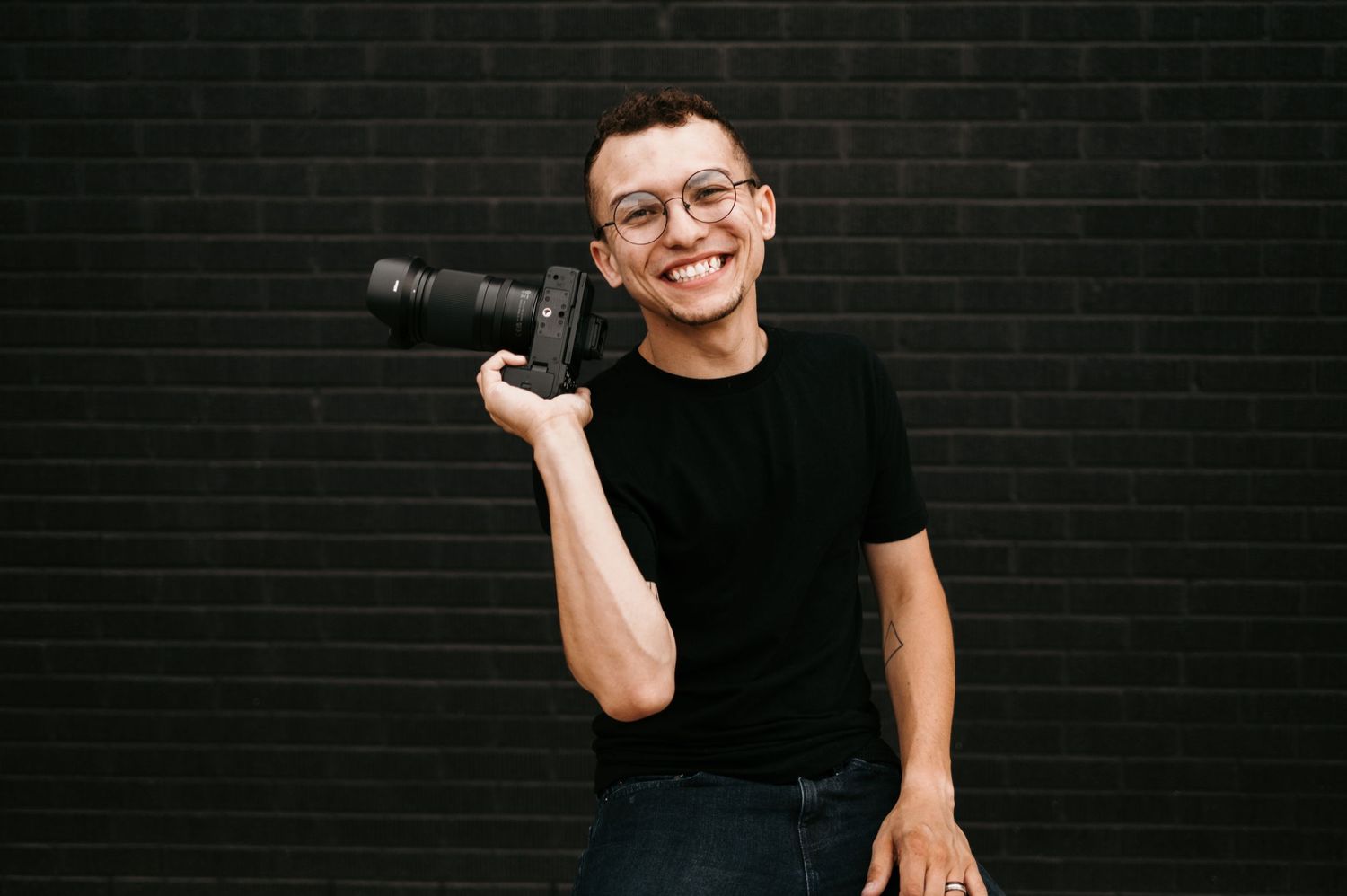 Austin Carter in a black t-shirt poses with camera equipment against a dark brick wall.
