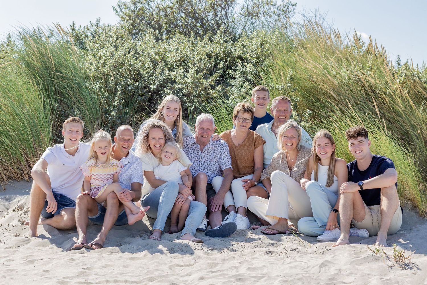Large family group smiling together on a sandy beach with green dunes, enjoying a sunny summer day outdoors.