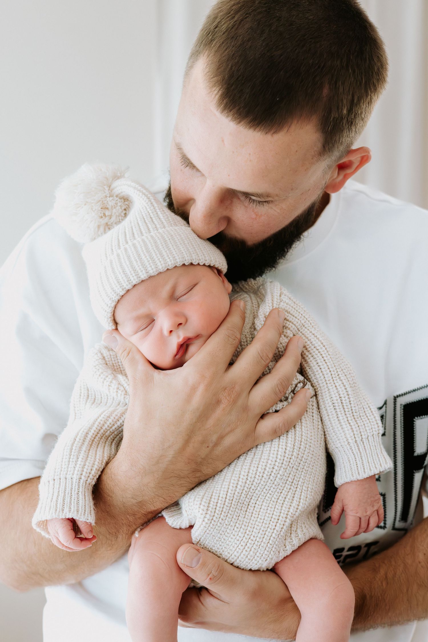 Bearded father tenderly kissing sleeping newborn baby dressed in knit hat and outfit.