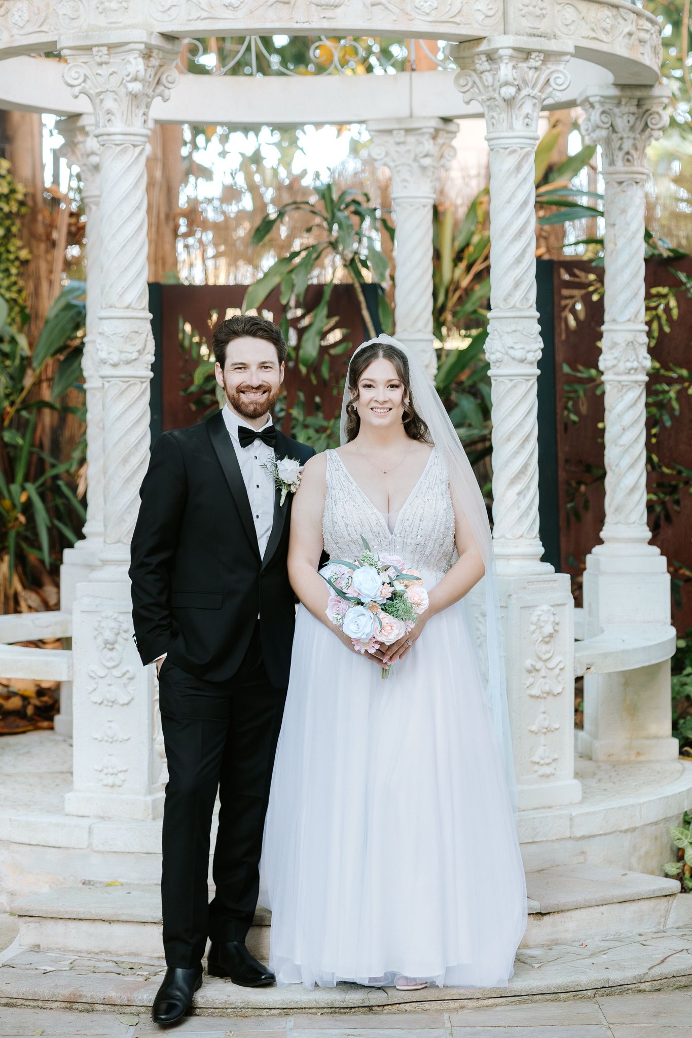 A bride and groom pose smiling under an ornate white gazebo, she holds a bouquet in a tulle gown, he wears a black tuxedo.