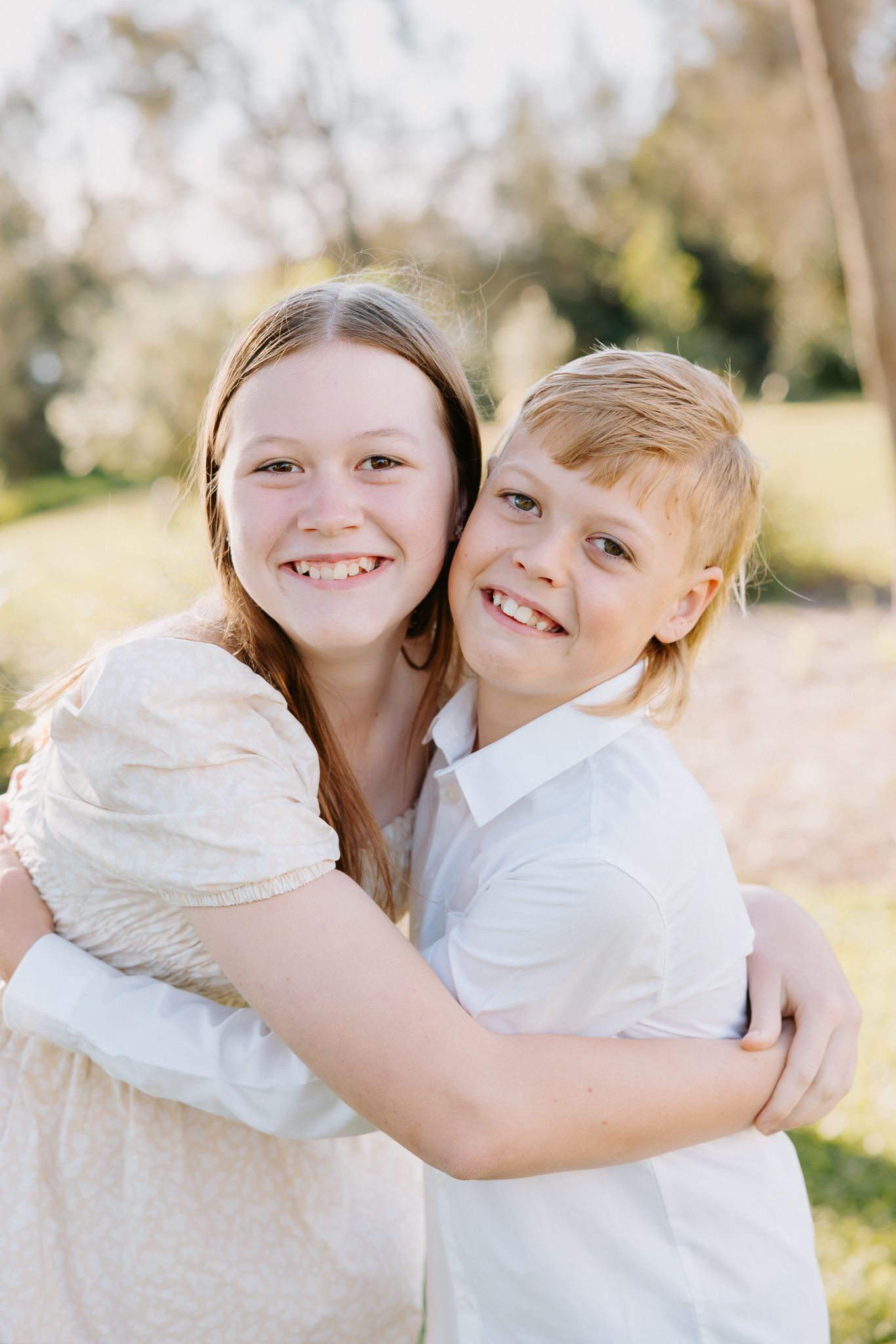Two smiling children hugging outdoors in a sunny park, a girl in a cream dress and a boy in a white shirt.