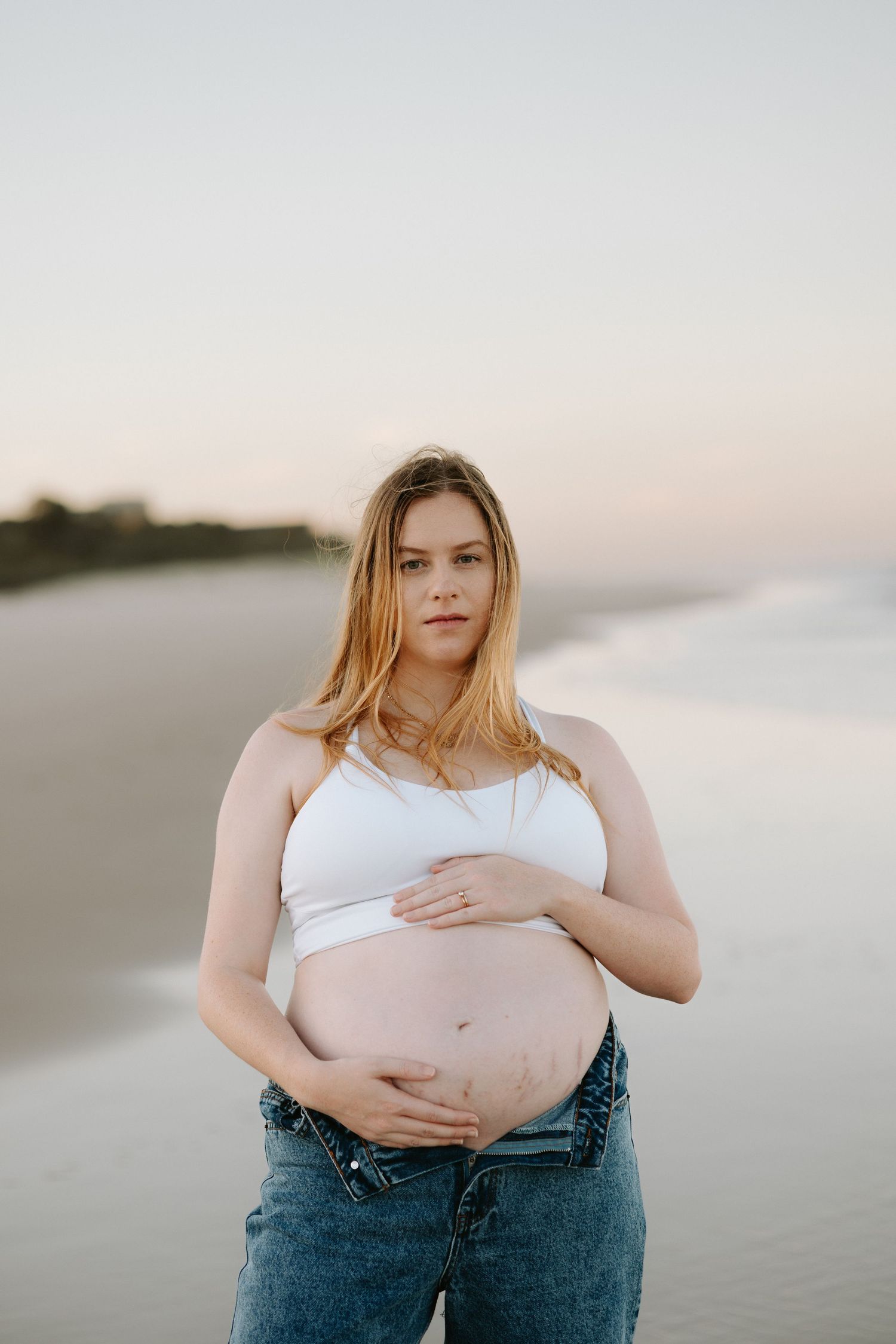 A series of maternity photos on a beach showing a pregnant woman in jeans and white top at sunset.
