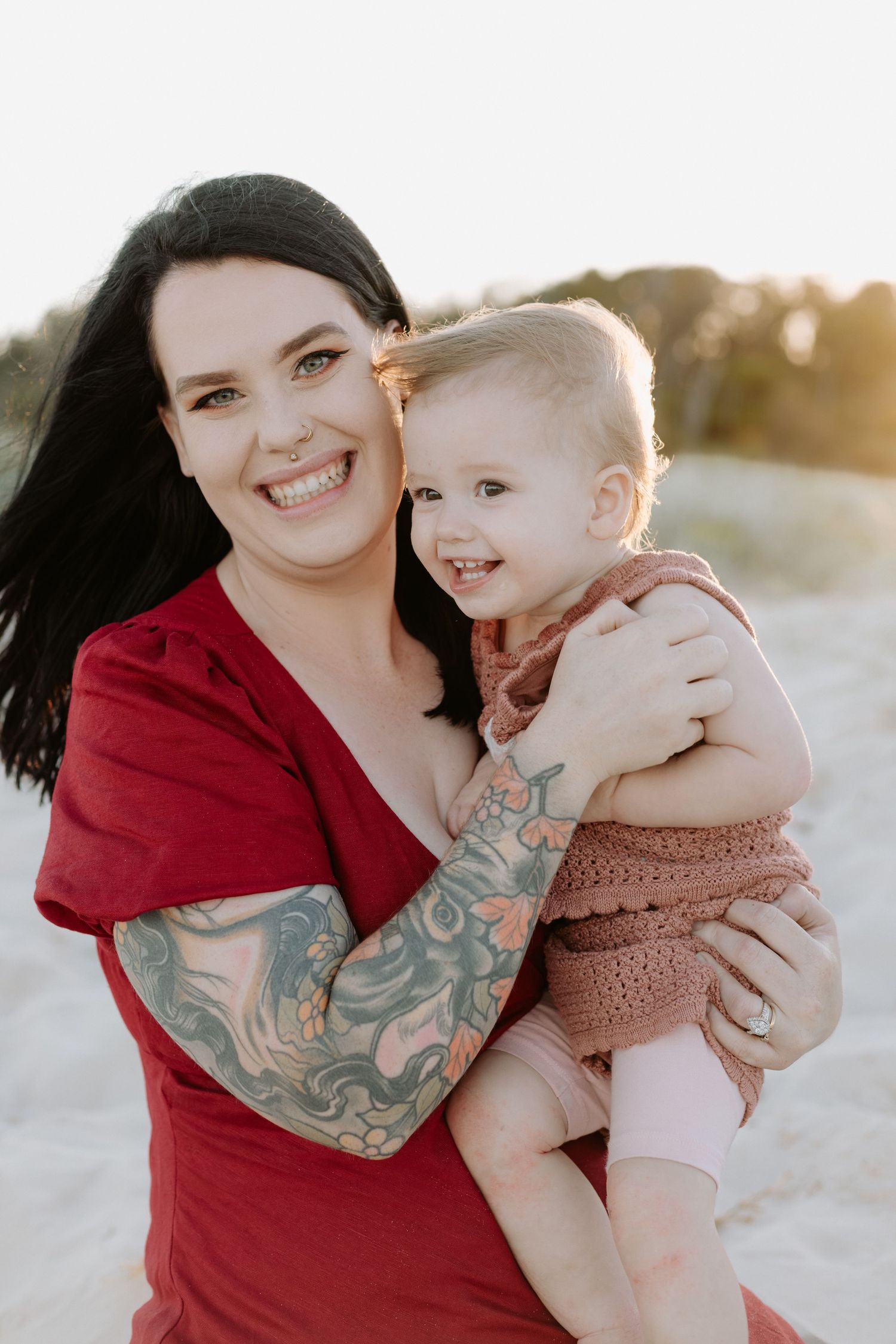 A parent in a red dress with sleeve tattoos holds a smiling toddler in a pink romper during golden hour outdoors.