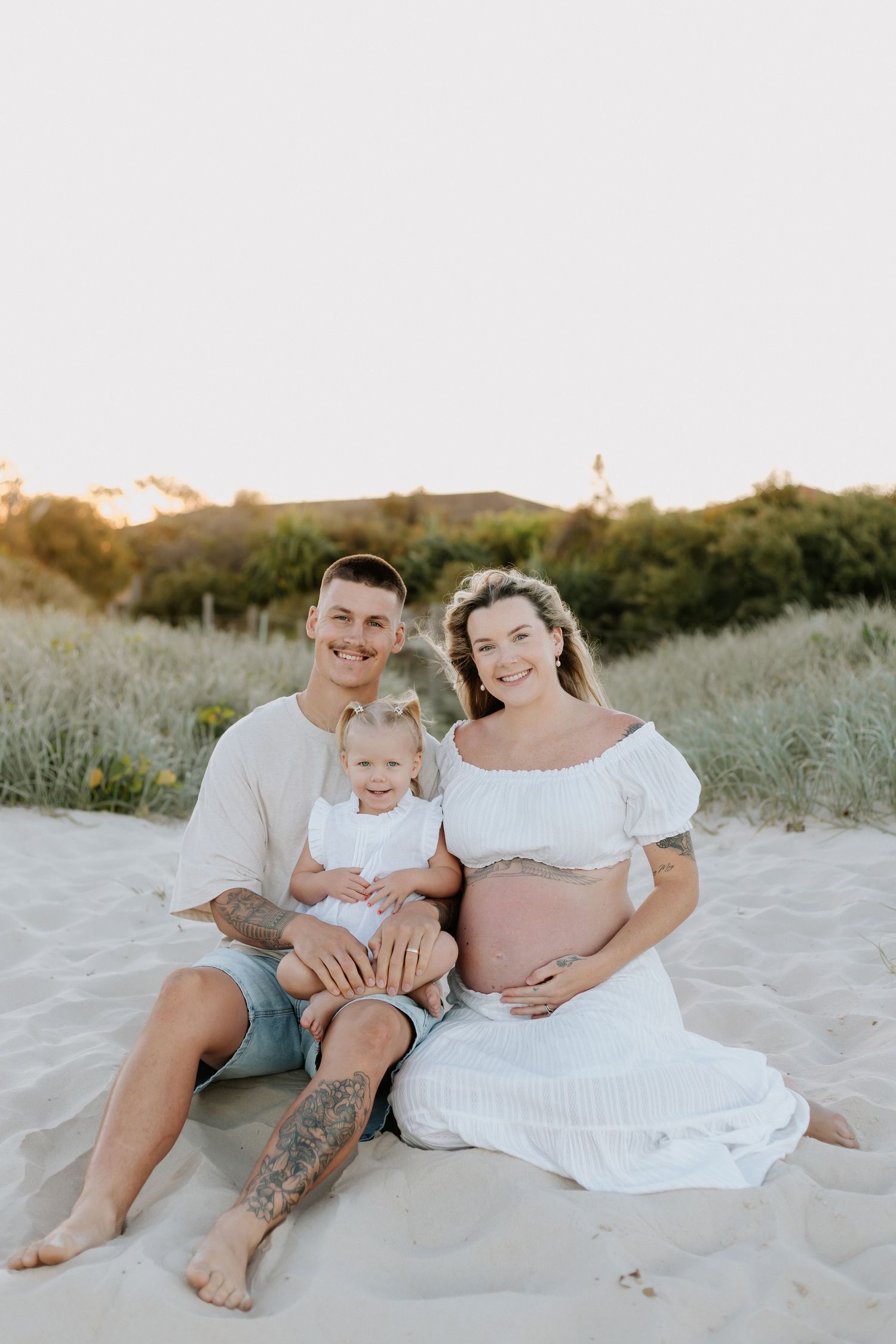 A family in light-colored clothing sits together on a sandy beach at sunset.