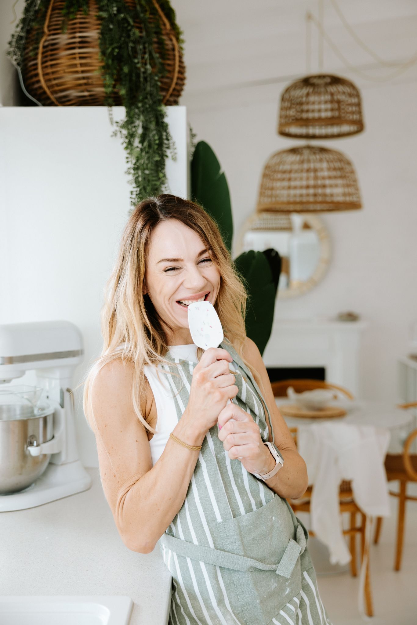 Woman in striped apron licking a white ice cream bar in a bright, modern kitchen with wicker pendant lights.