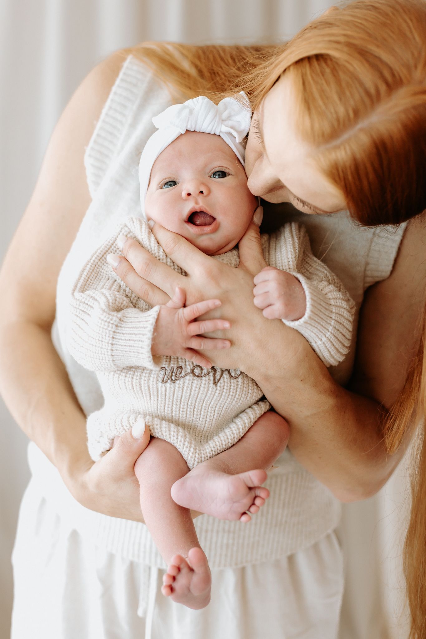 Red-haired mother tenderly holds her newborn baby wearing a white bow headband and beige knit onesie.