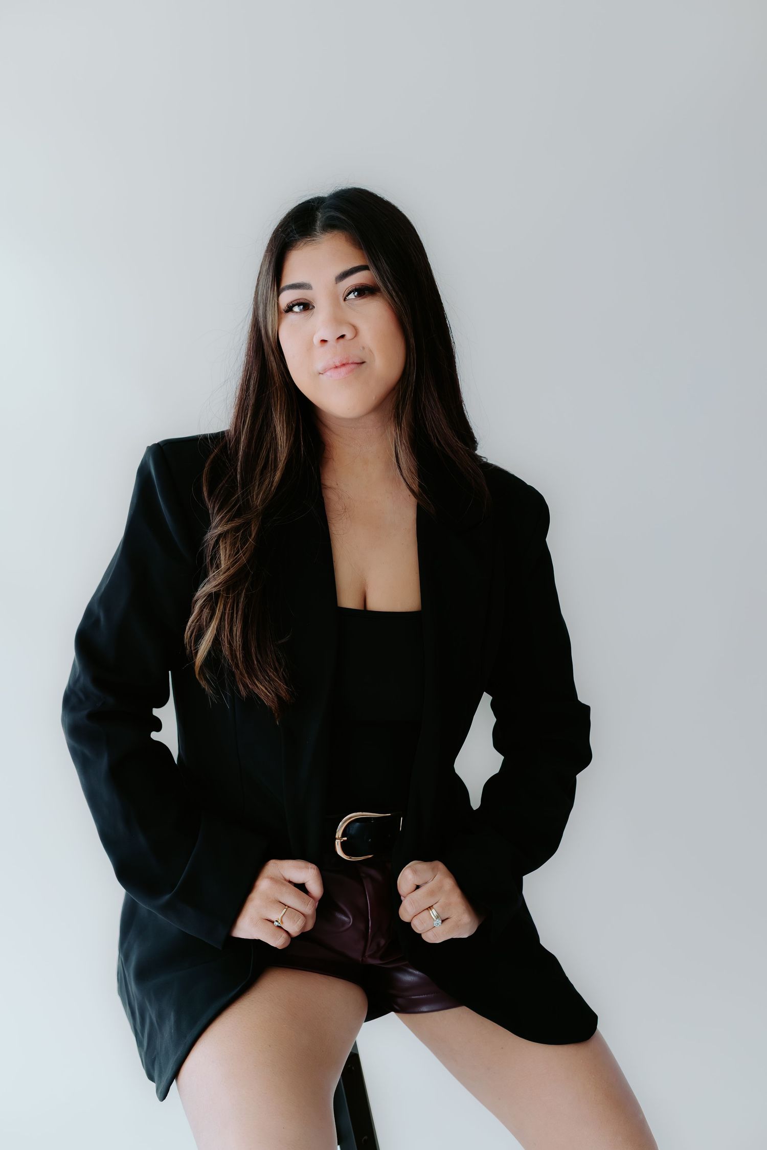 Woman in black blazer and shorts with belt poses confidently against white background.