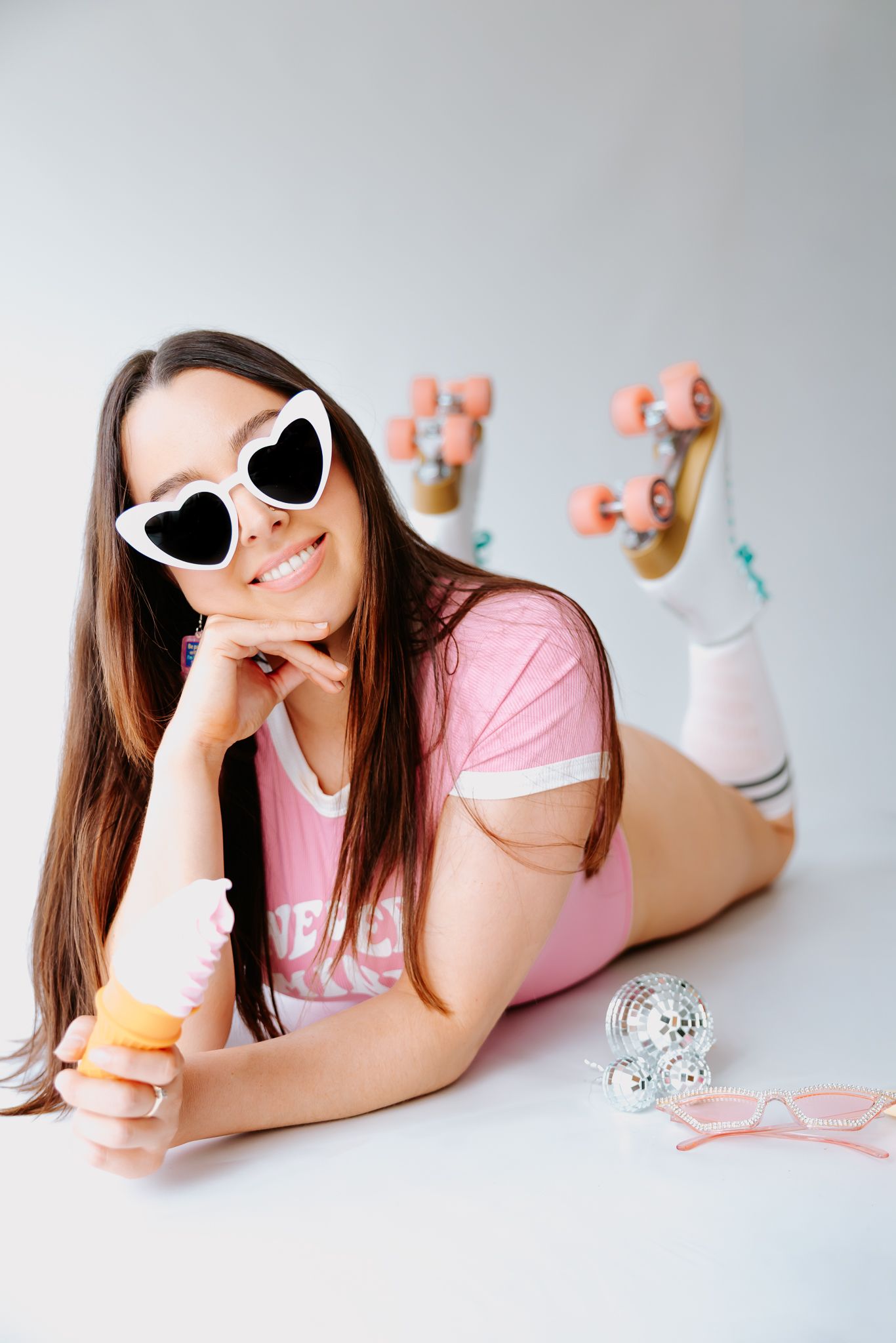 Young woman in pink outfit and heart sunglasses lying down with roller skates, holding a macaron on white background.