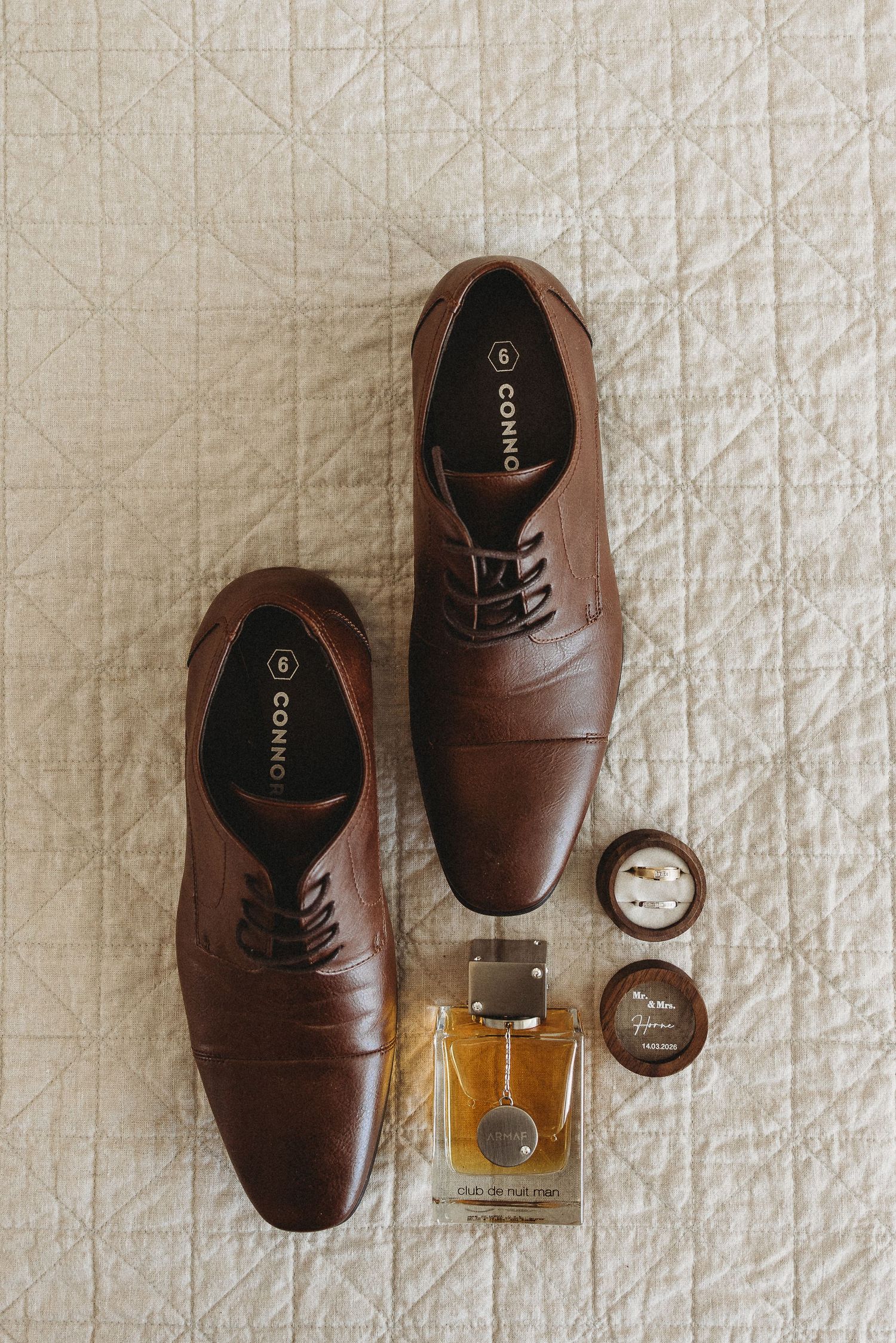 Brown leather Oxford dress shoes by Connо displayed on bed alongside cologne bottle and cufflinks for groom.