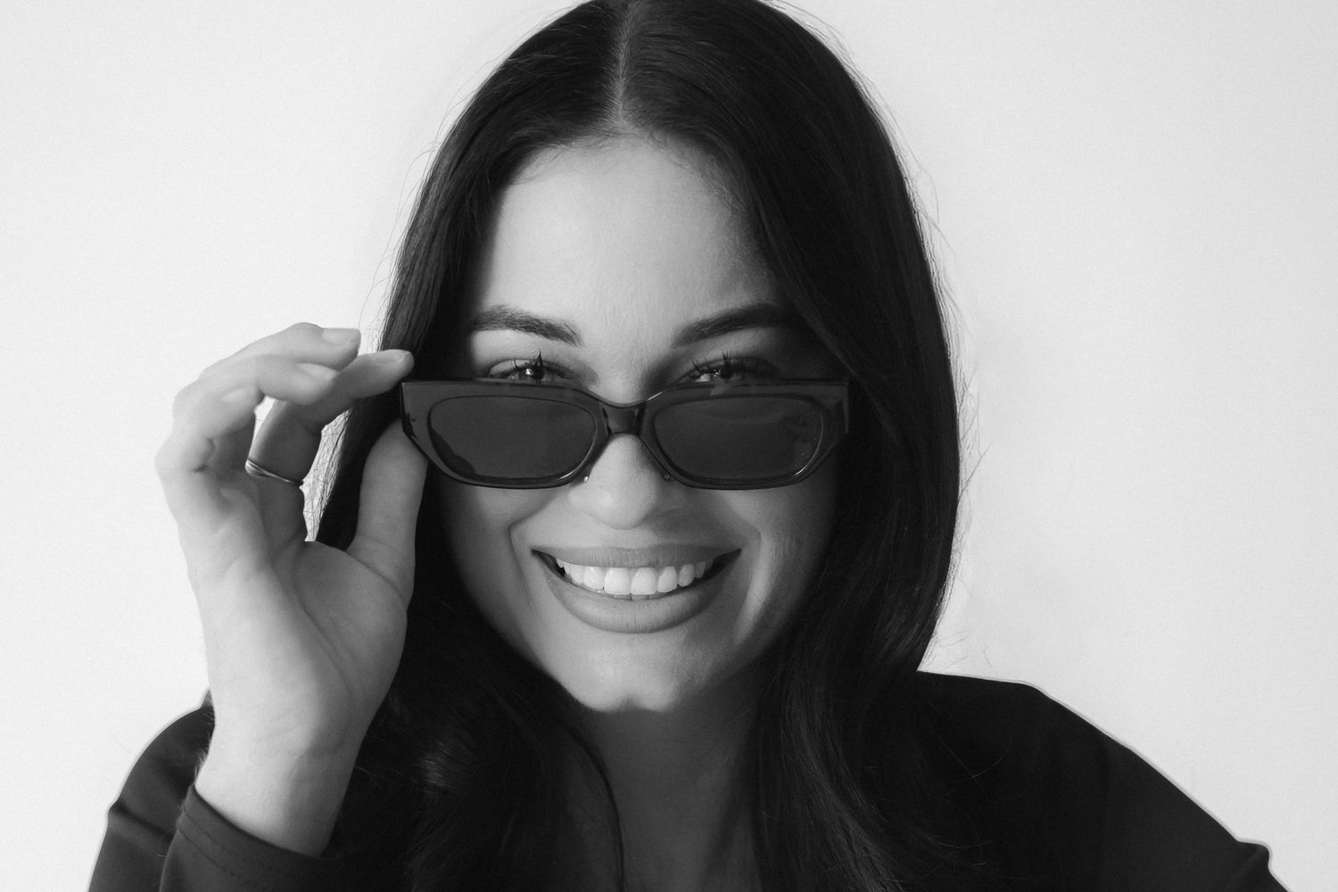 Black and white portrait of a smiling woman with long dark hair pulling down cat-eye sunglasses.