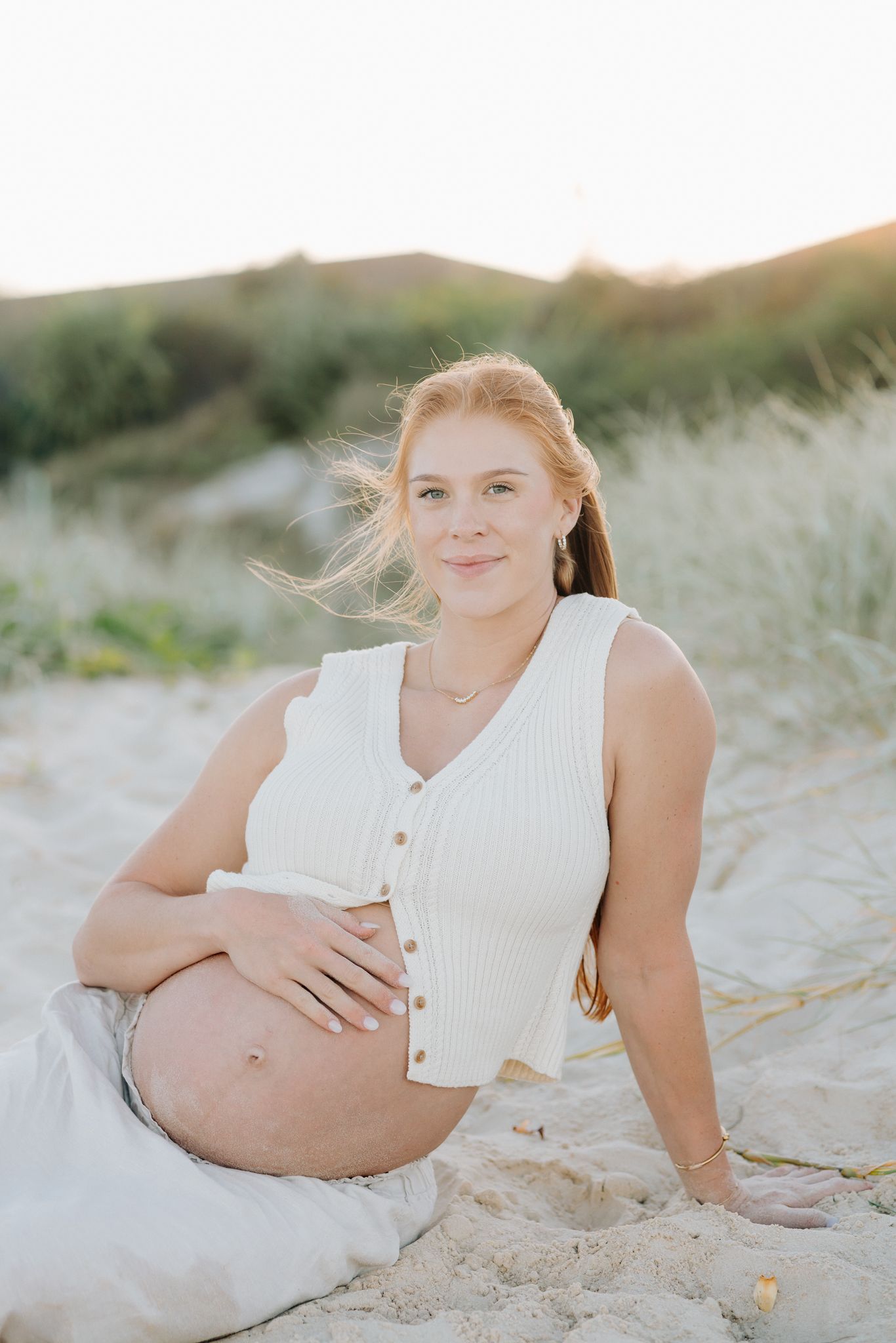 Pregnant woman sitting on sandy beach dunes at golden hour, cradling baby bump in white ribbed top.