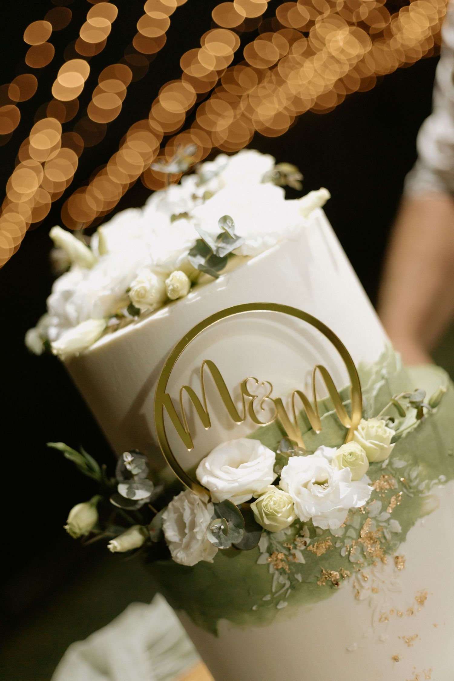 Elegant two-tier wedding cake with white roses, greenery, and gold M&M monogram topper, surrounded by bokeh fairy lights.