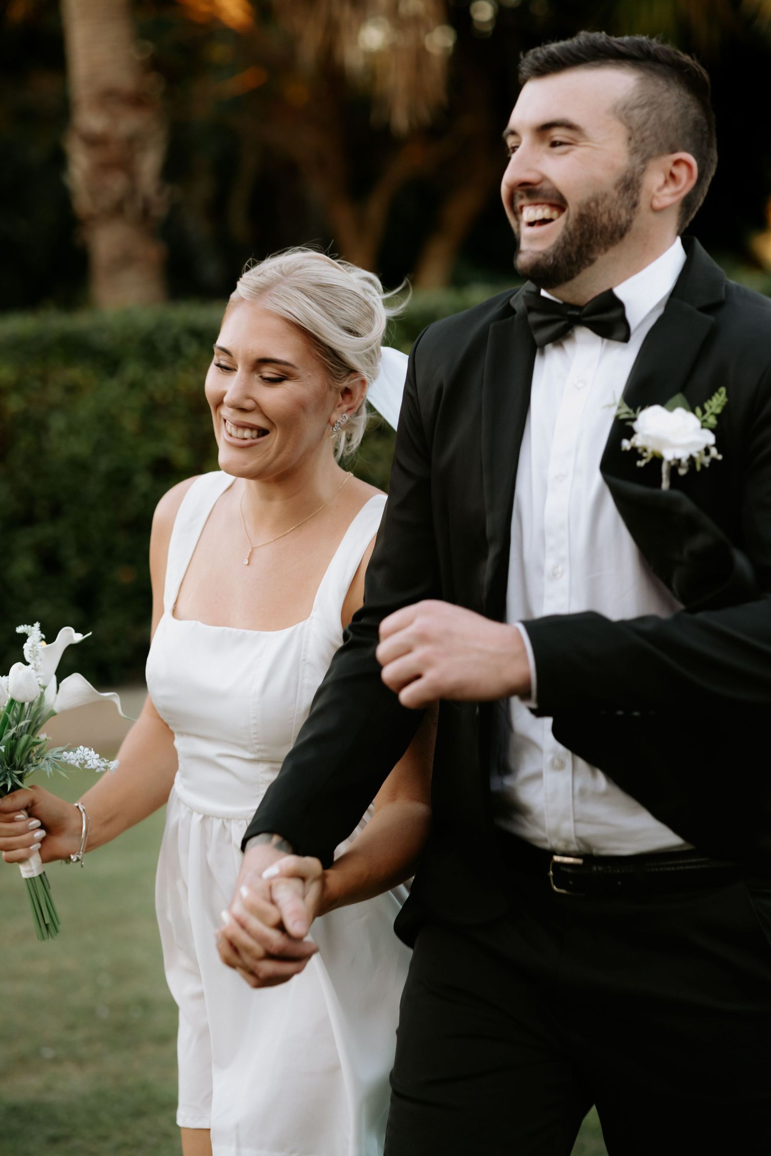 Happy bride in white dress holding bouquet walks arm-in-arm with groom in black tuxedo outdoors.