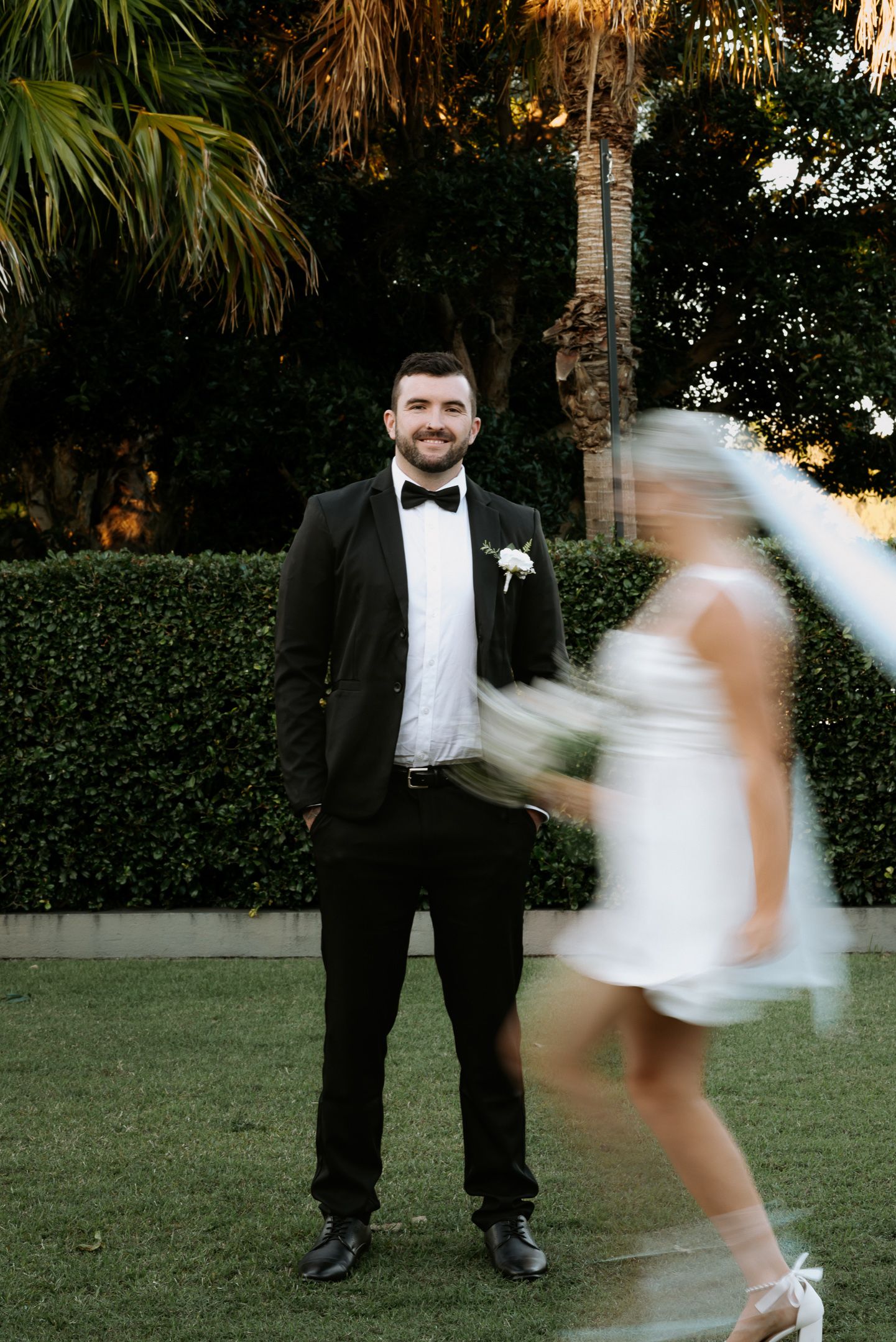 Groom in black tuxedo smiles as blurred bride in short white dress walks past on green lawn.