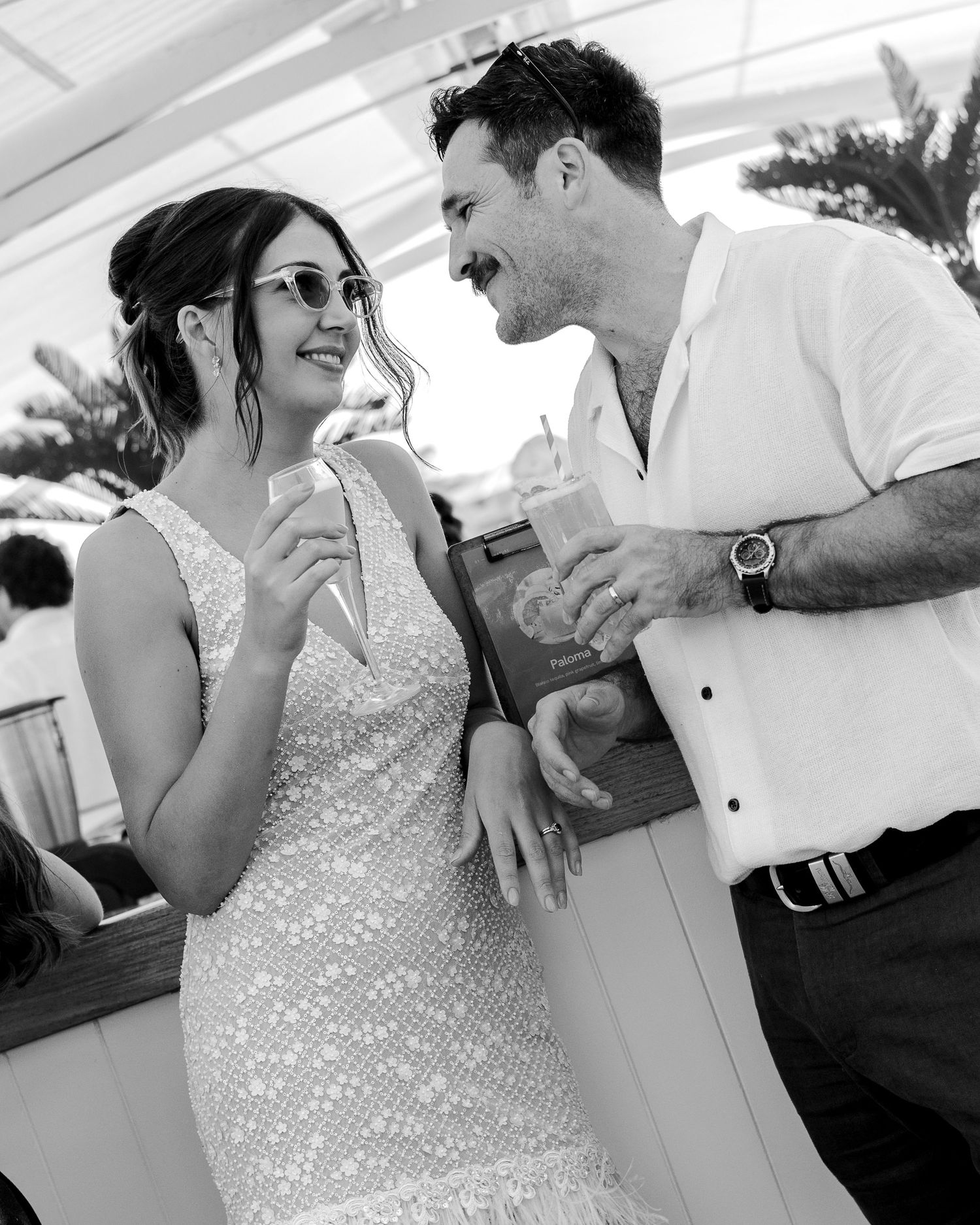 Black and white photo of a smiling couple in formal attire holding drinks at an outdoor wedding reception.