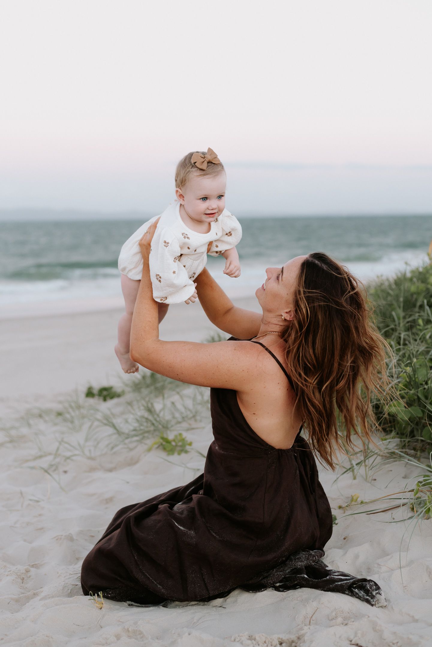 A mother in a black dress lifts and plays with a baby wearing white while sitting on a sandy beach at sunset.