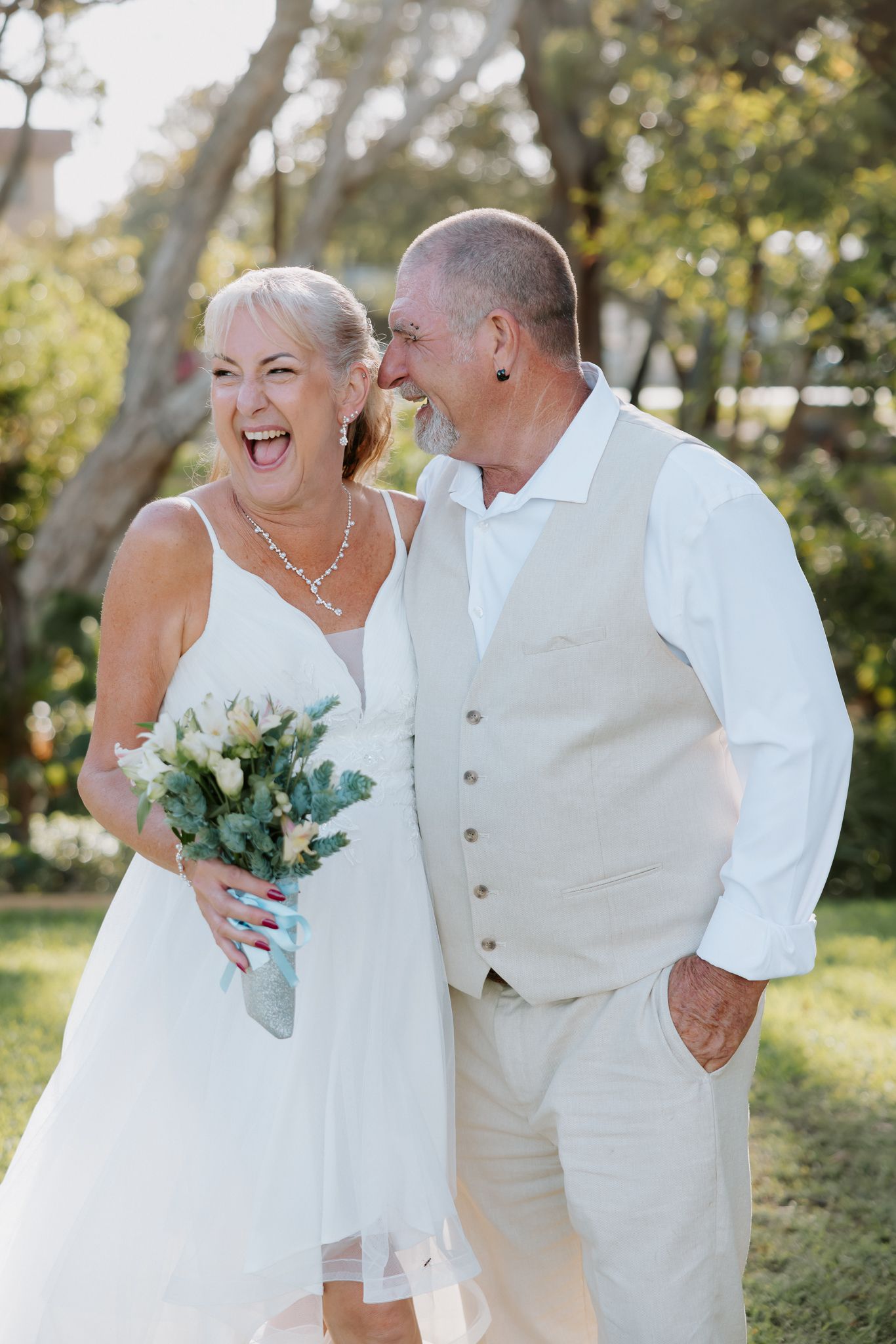 Joyful mature couple in white wedding attire laughing outdoors, bride holding bouquet of white flowers.
