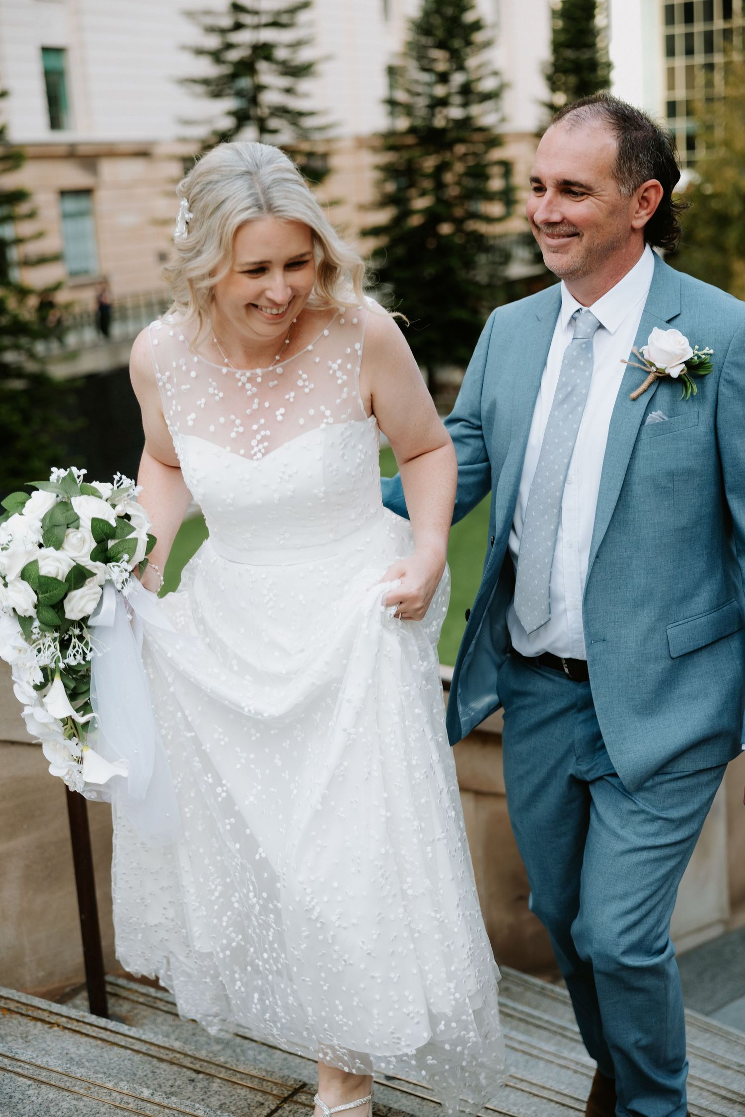 A couple in wedding attire share candid moments on steps, with the bride in a white lace dress and groom in blue suit.