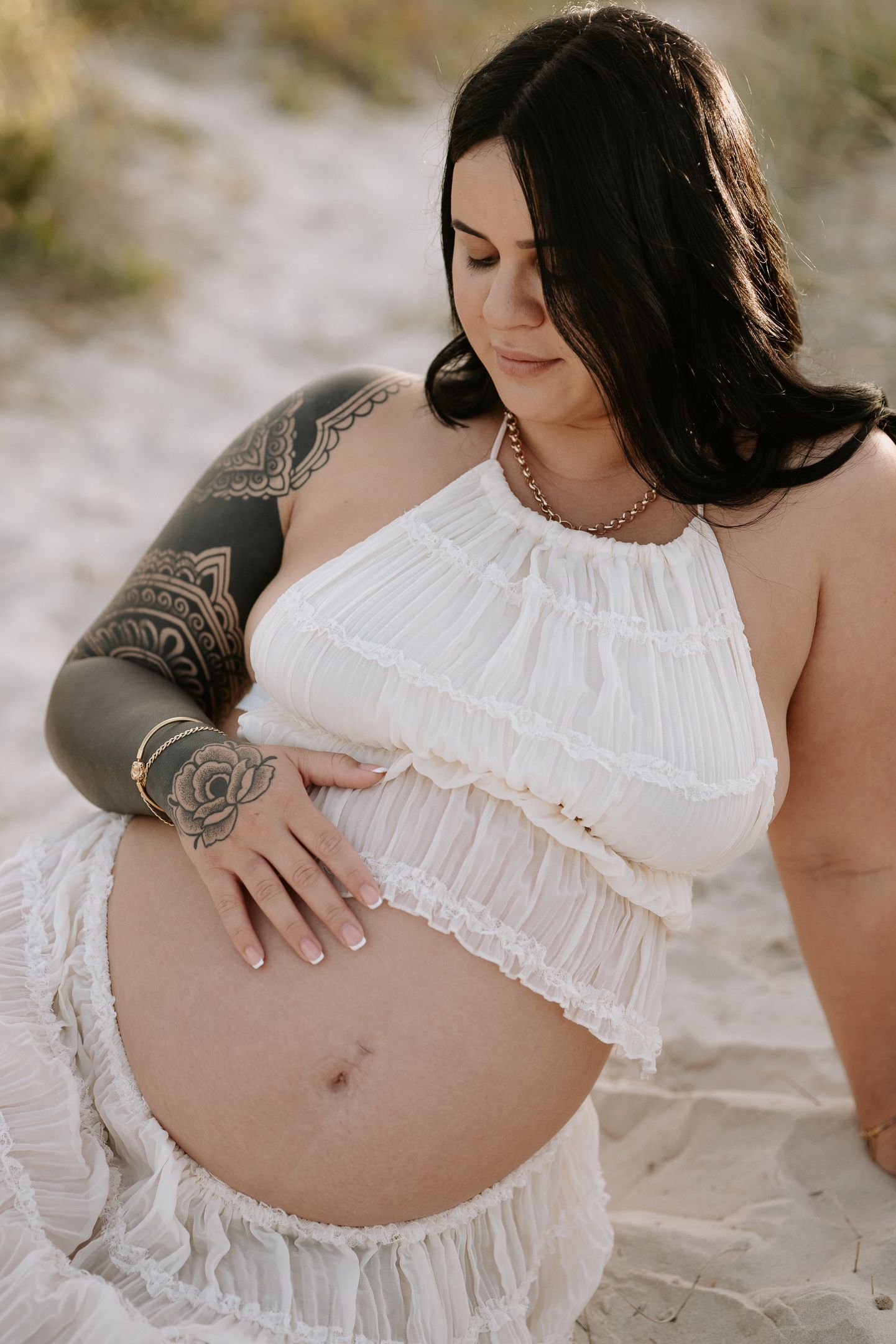 Pregnant woman in white flowing dress poses on beach showing baby bump with ornate arm tattoo sleeve in sunlit setting.