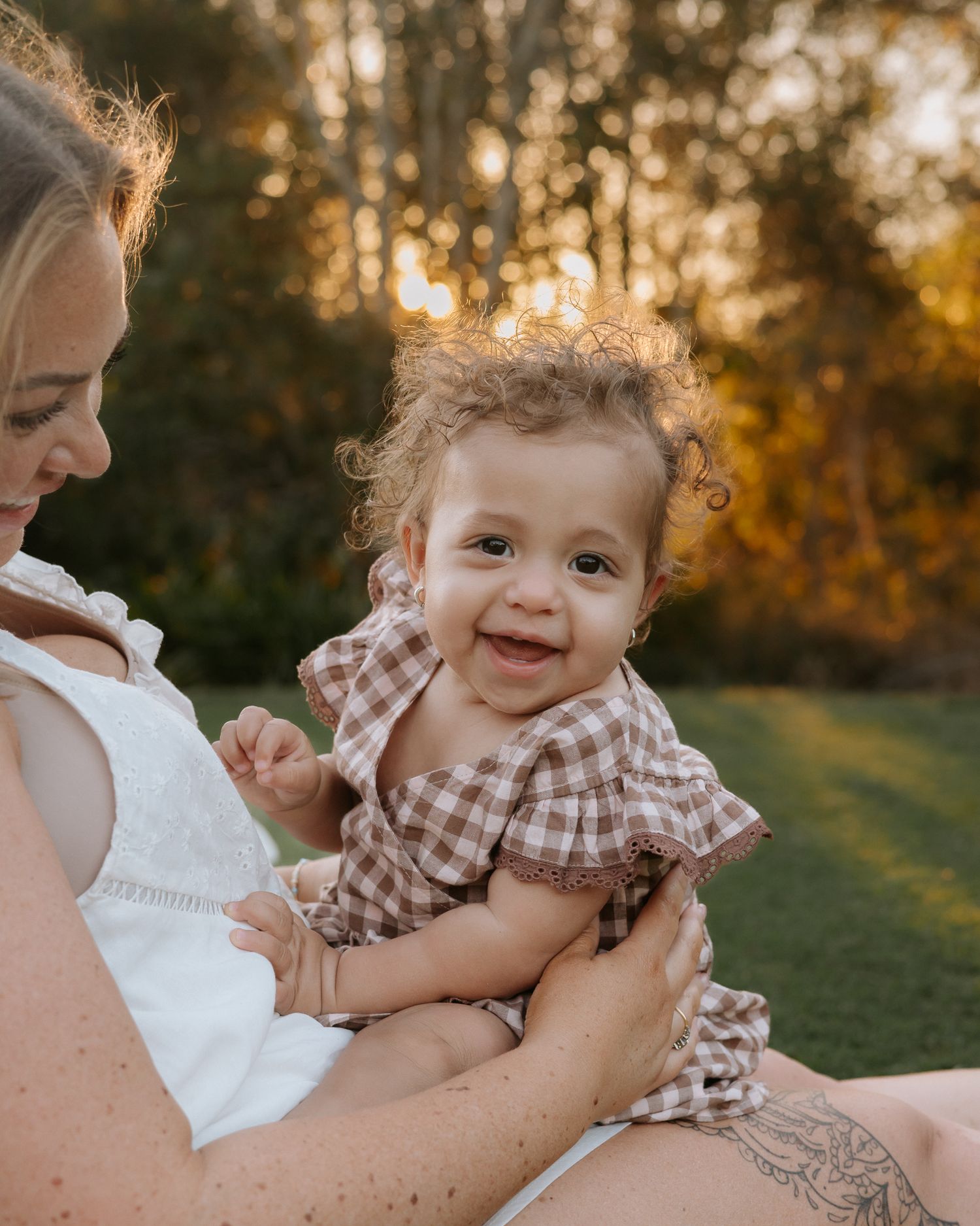 A smiling baby in a gingham dress being held by a mother outdoors during golden hour.