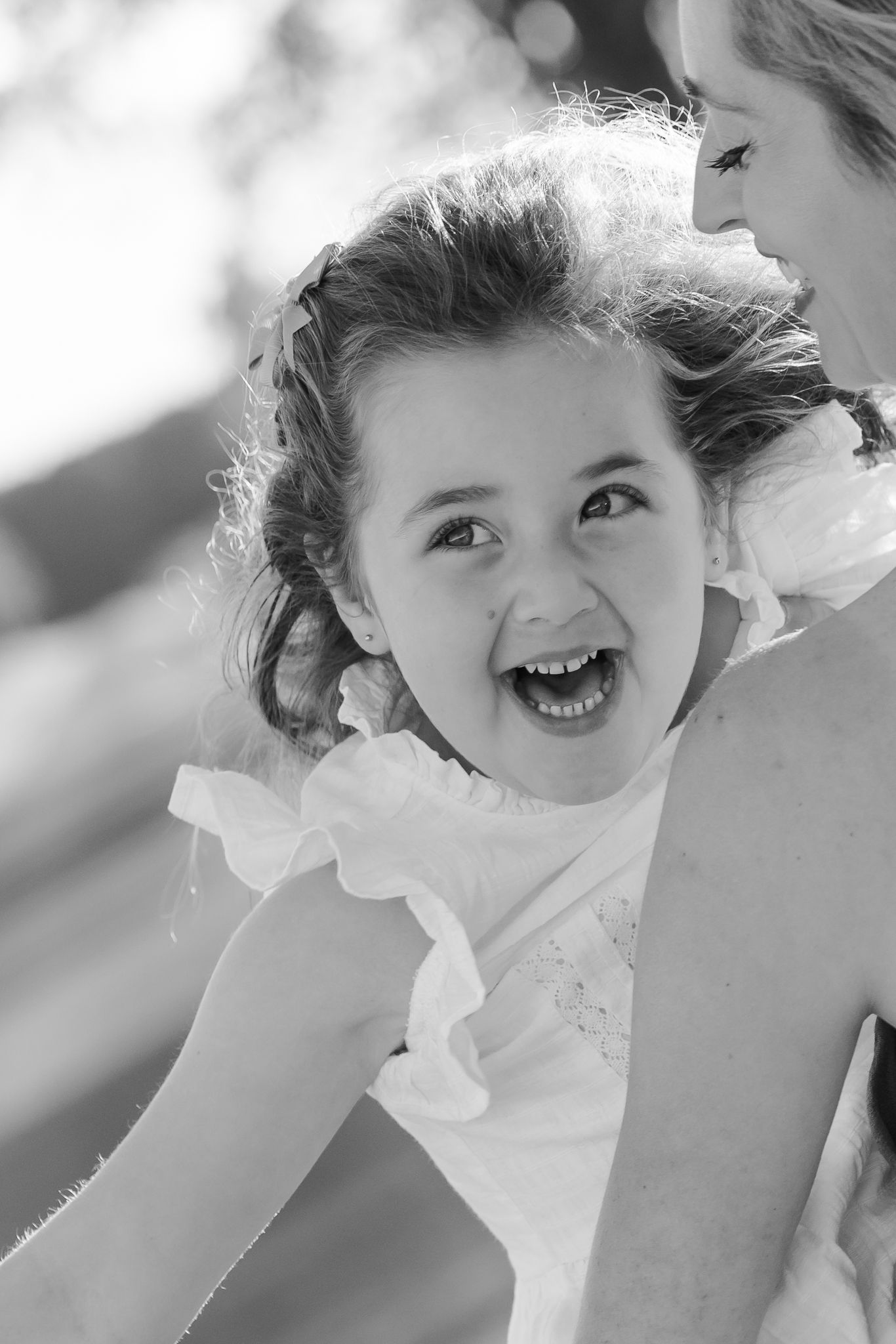Black and white photo of a joyful young girl in a white dress laughing while being held by an adult outdoors.