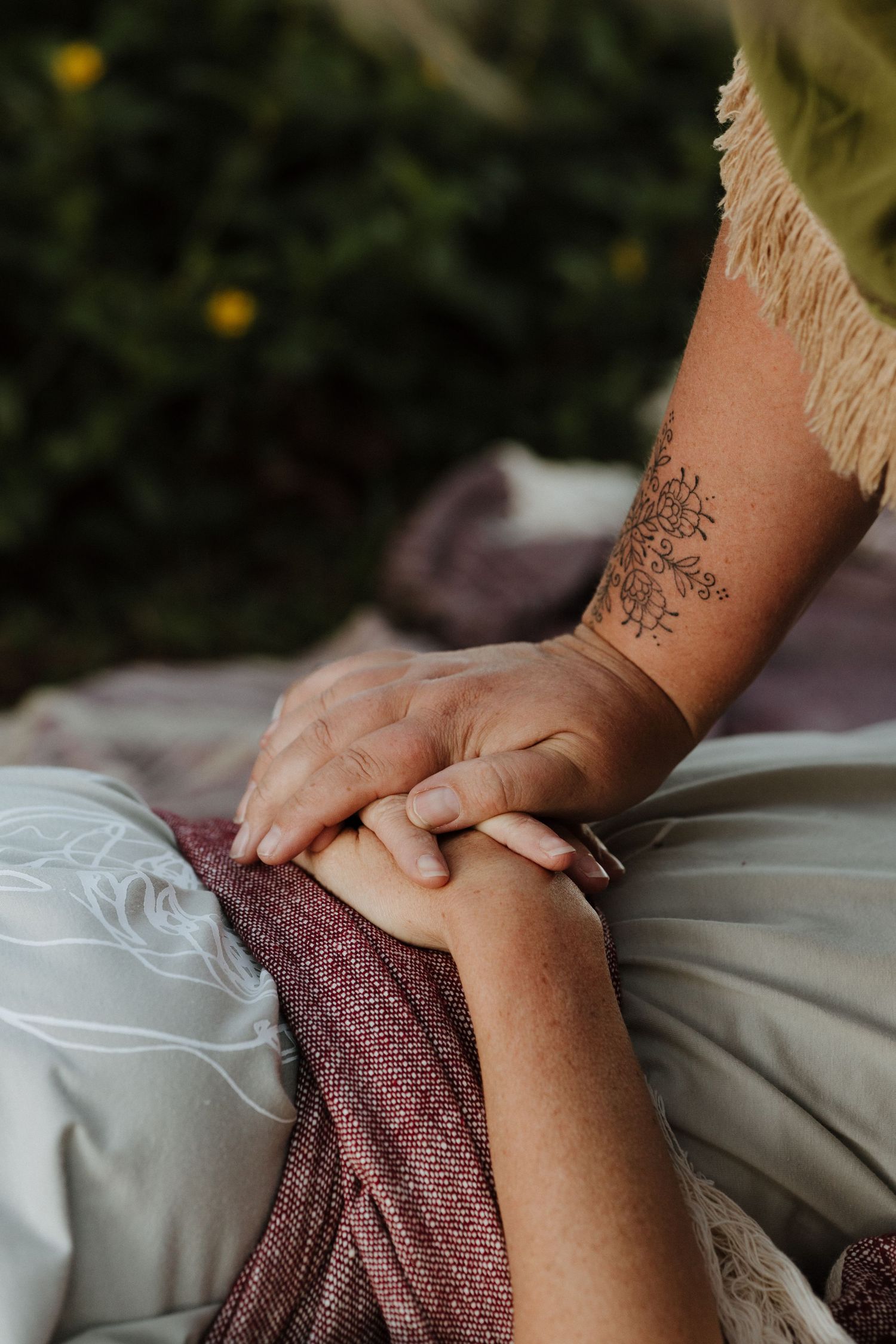 Close-up of two people holding hands outdoors, one with a floral wrist tattoo, sitting on soft blankets.