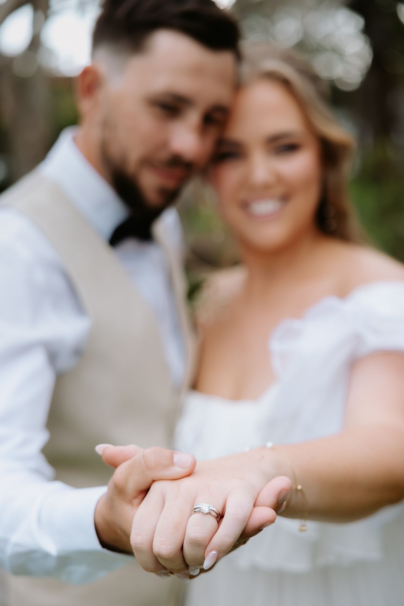 Newlywed couple displaying their wedding rings, bride in off-shoulder white dress smiling at outdoor ceremony.