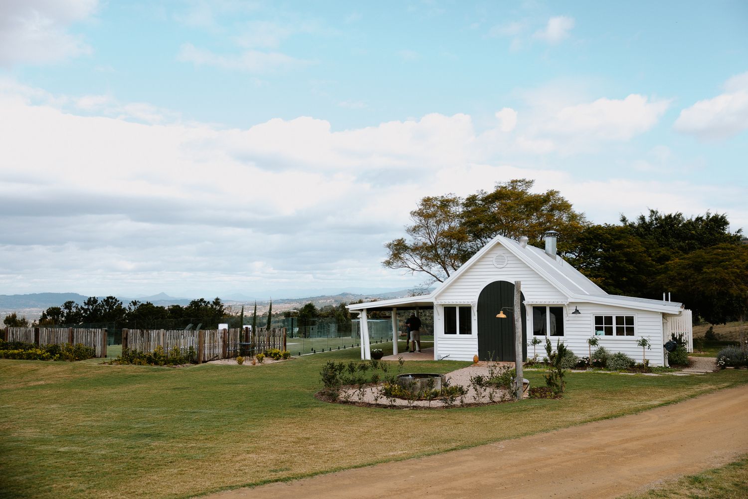White country farmhouse with green lawn, circular garden bed, and wooden fence under a blue sky.