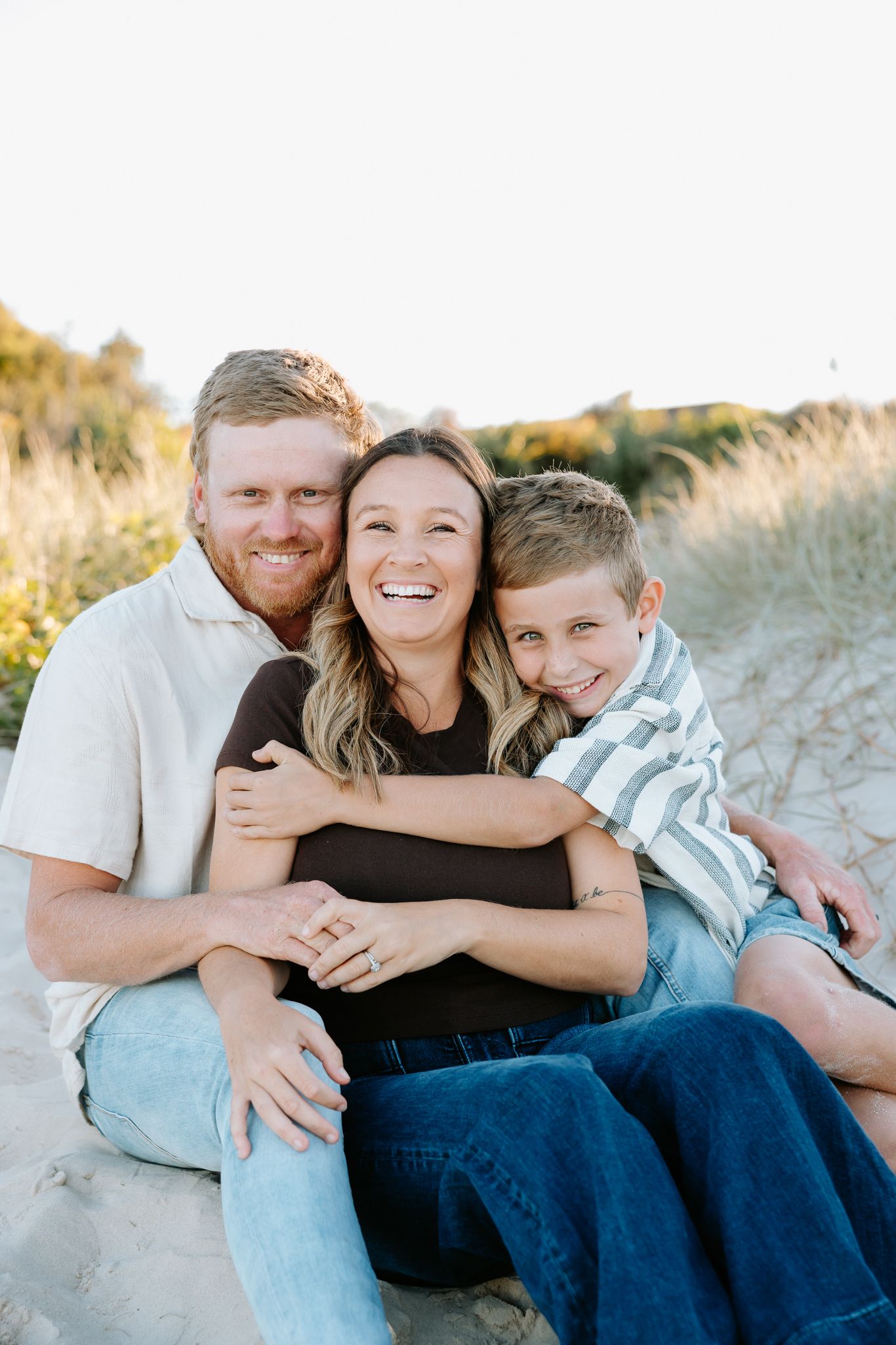 Happy family of three smiling together on sandy beach dunes, parents and young boy posing for portrait.