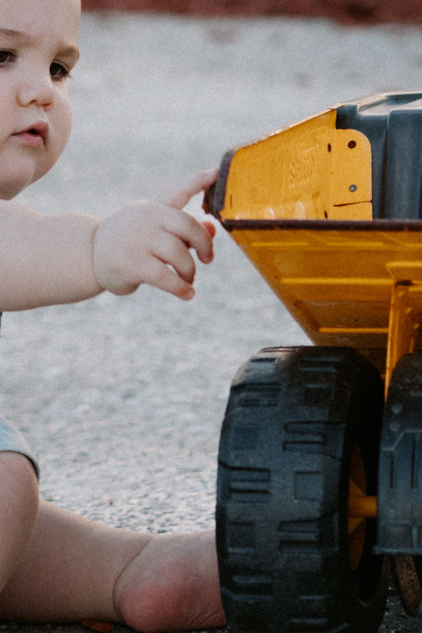 A baby plays with a large yellow toy dump truck outdoors on a gravel surface.