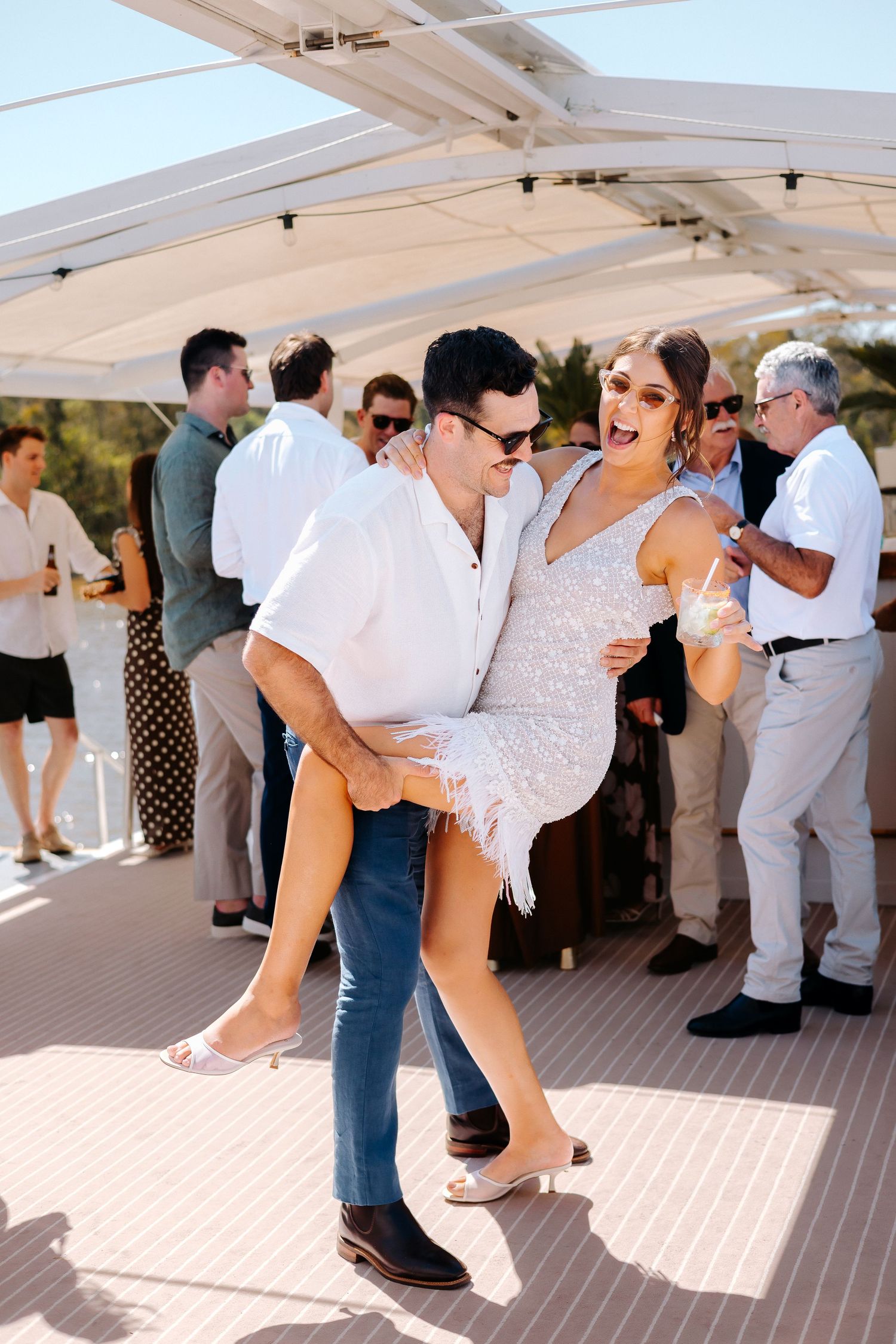 Couple dancing joyfully at an outdoor party, woman in white sequin dress lifted by man in white shirt and jeans.