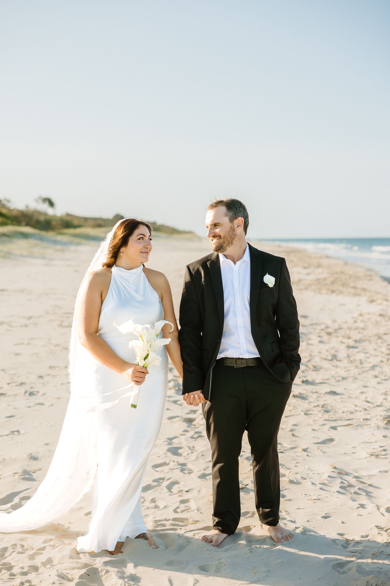 Bride and groom walking barefoot on a sandy beach, holding hands and smiling at each other in wedding attire.