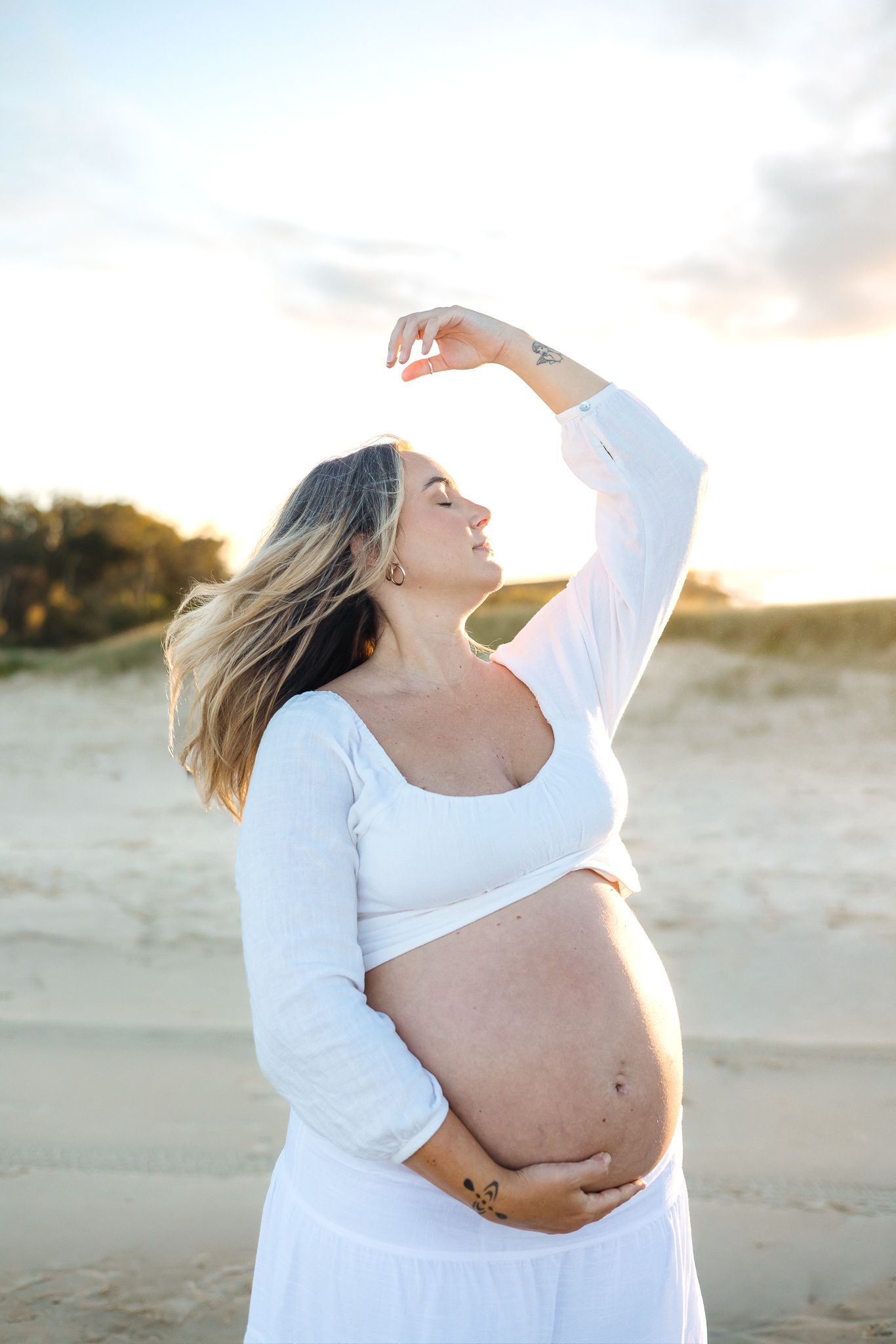 Pregnant woman in white crop top and skirt cradling baby bump on sandy beach at golden hour sunset.