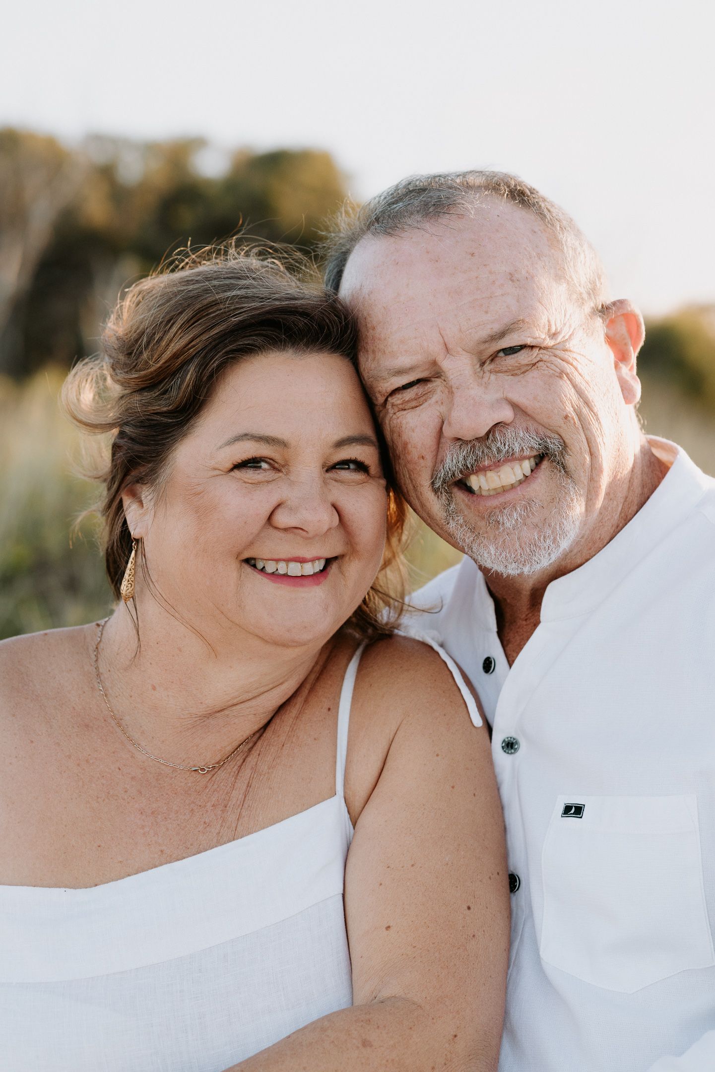 Happy middle-aged couple smiling cheek to cheek outdoors, wearing white, with golden hour sunlight in background.