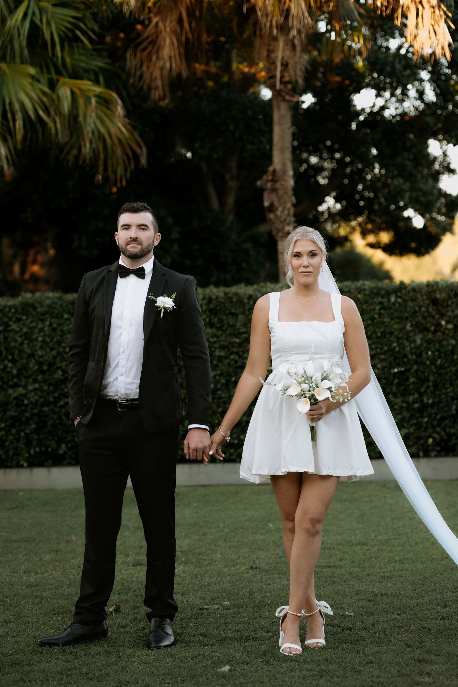 Bride in short white dress holding bouquet stands with groom in black tuxedo on green lawn with palm trees.