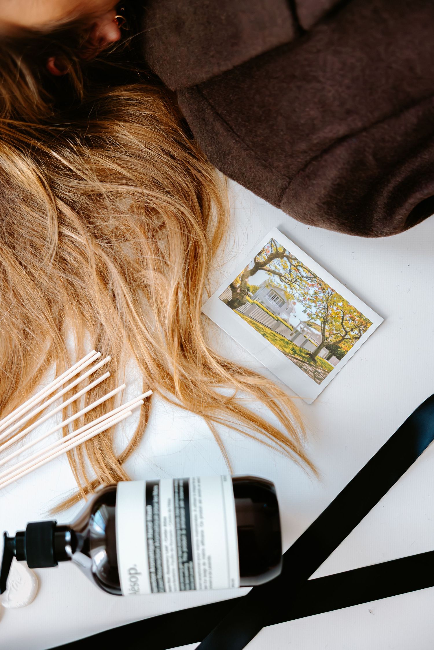 Flatlay of blonde hair, Aesop bottle, reed diffuser sticks, a small photo print, and black ribbon on white surface.