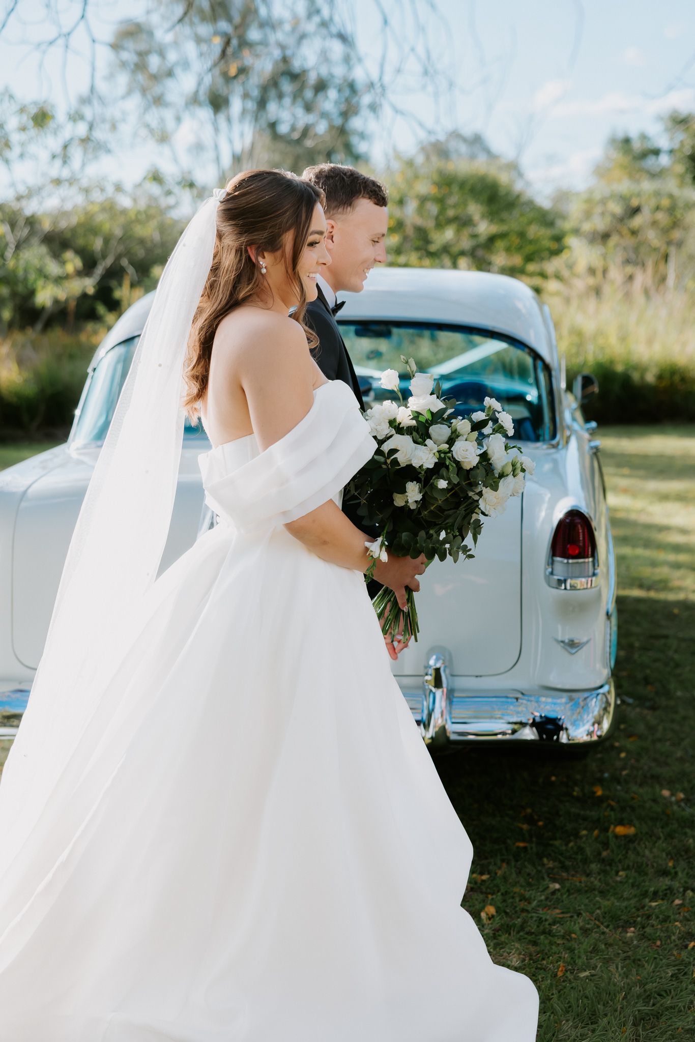 Bride in off-shoulder white gown with veil holds bouquet beside groom near vintage white car outdoors.