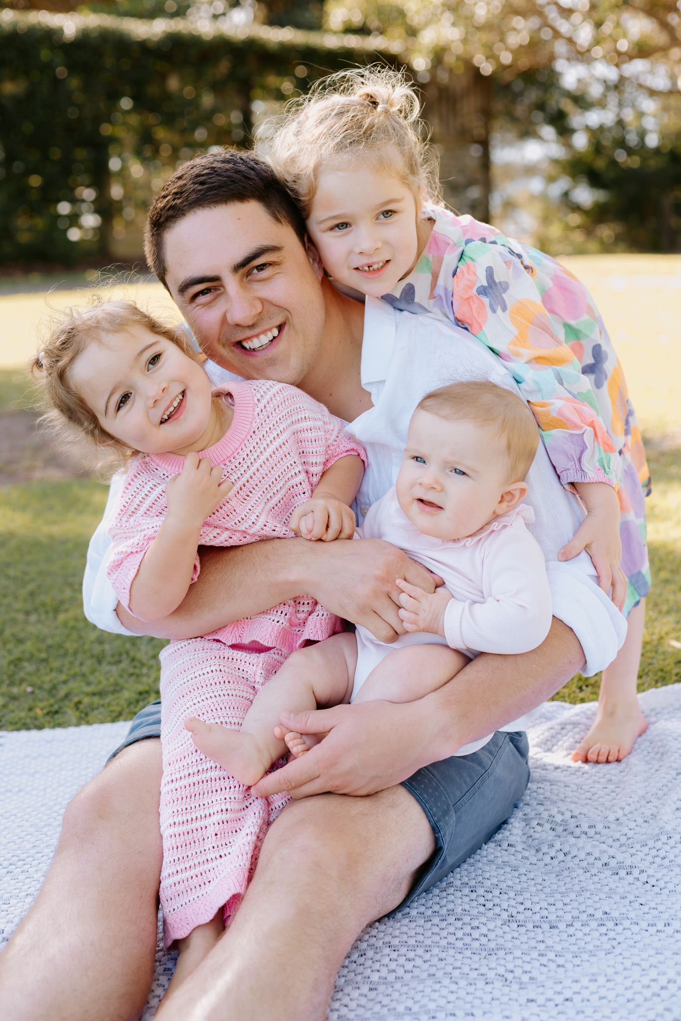 A smiling father sits on a blanket outdoors, hugging his three young children, including two girls and a baby.