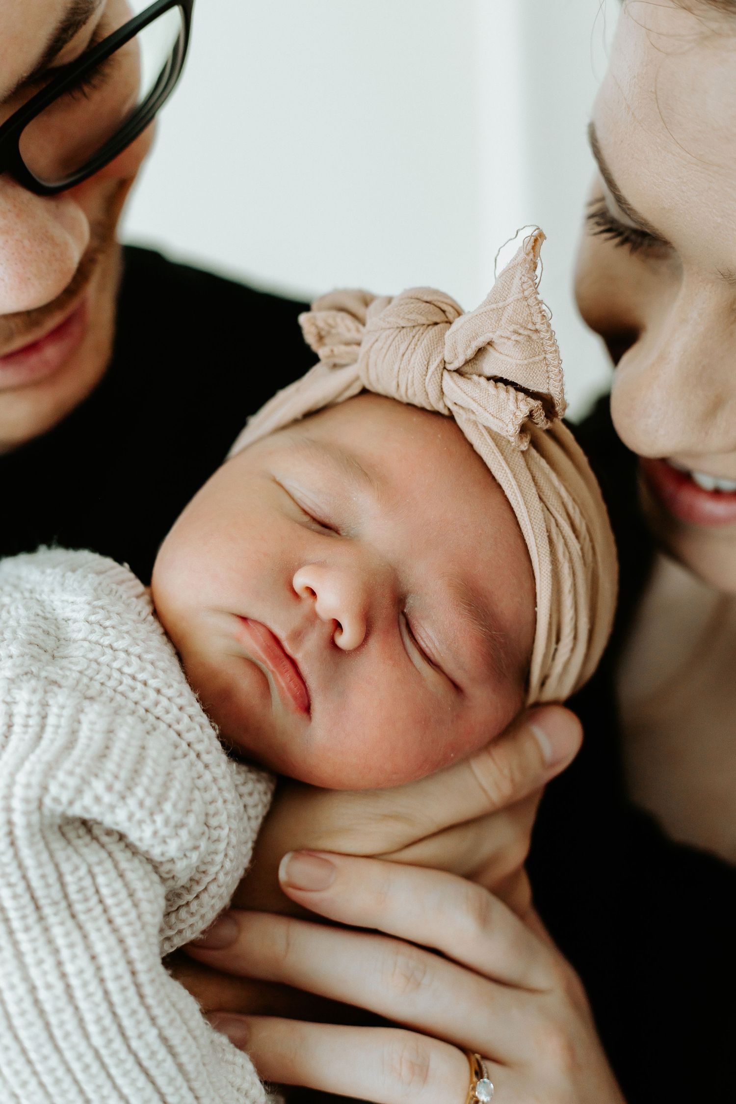 Two parents lovingly cradling their sleeping newborn baby girl wearing a beige bow headband.