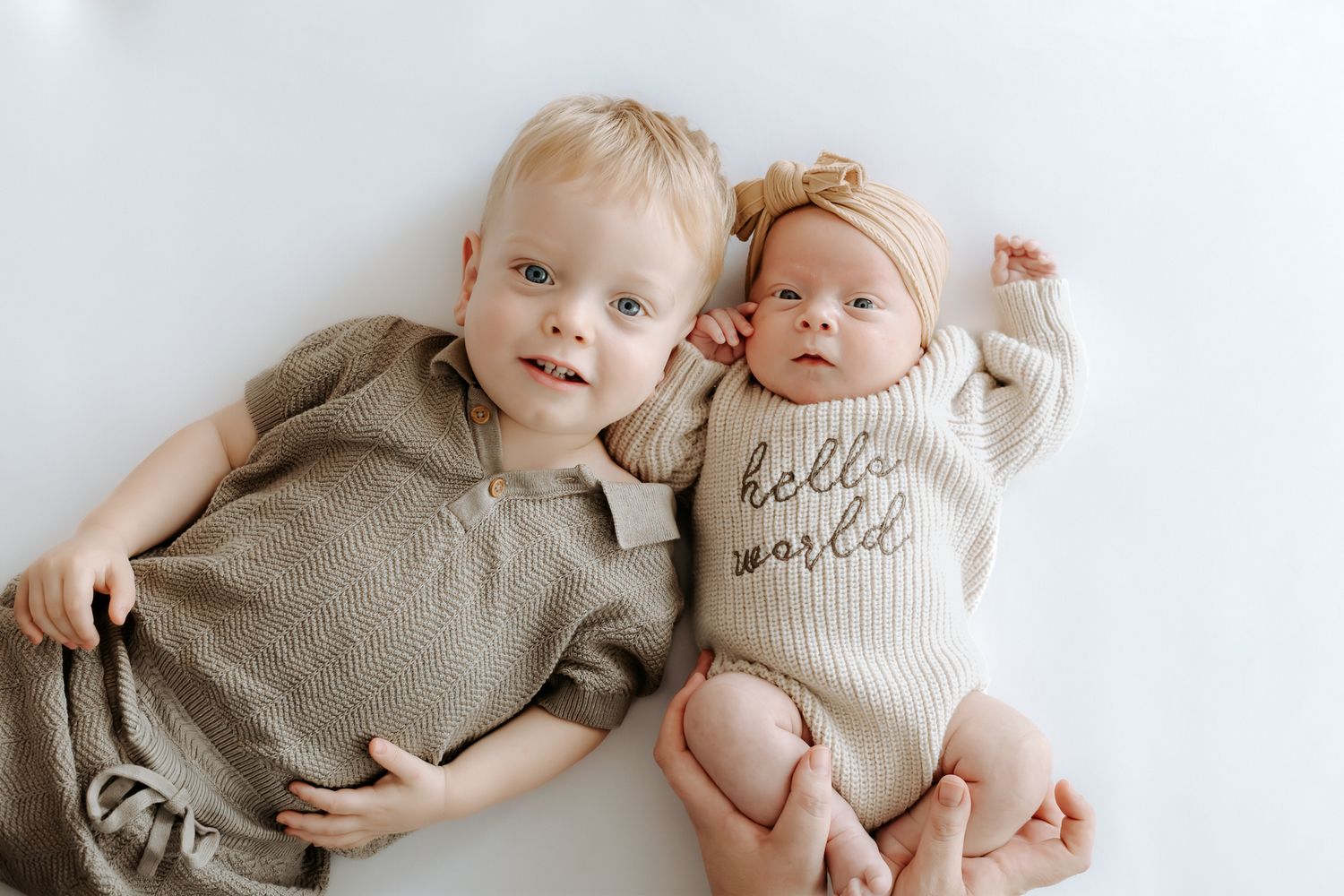 Toddler boy and newborn baby in 'hello world' onesie lying together on white background.